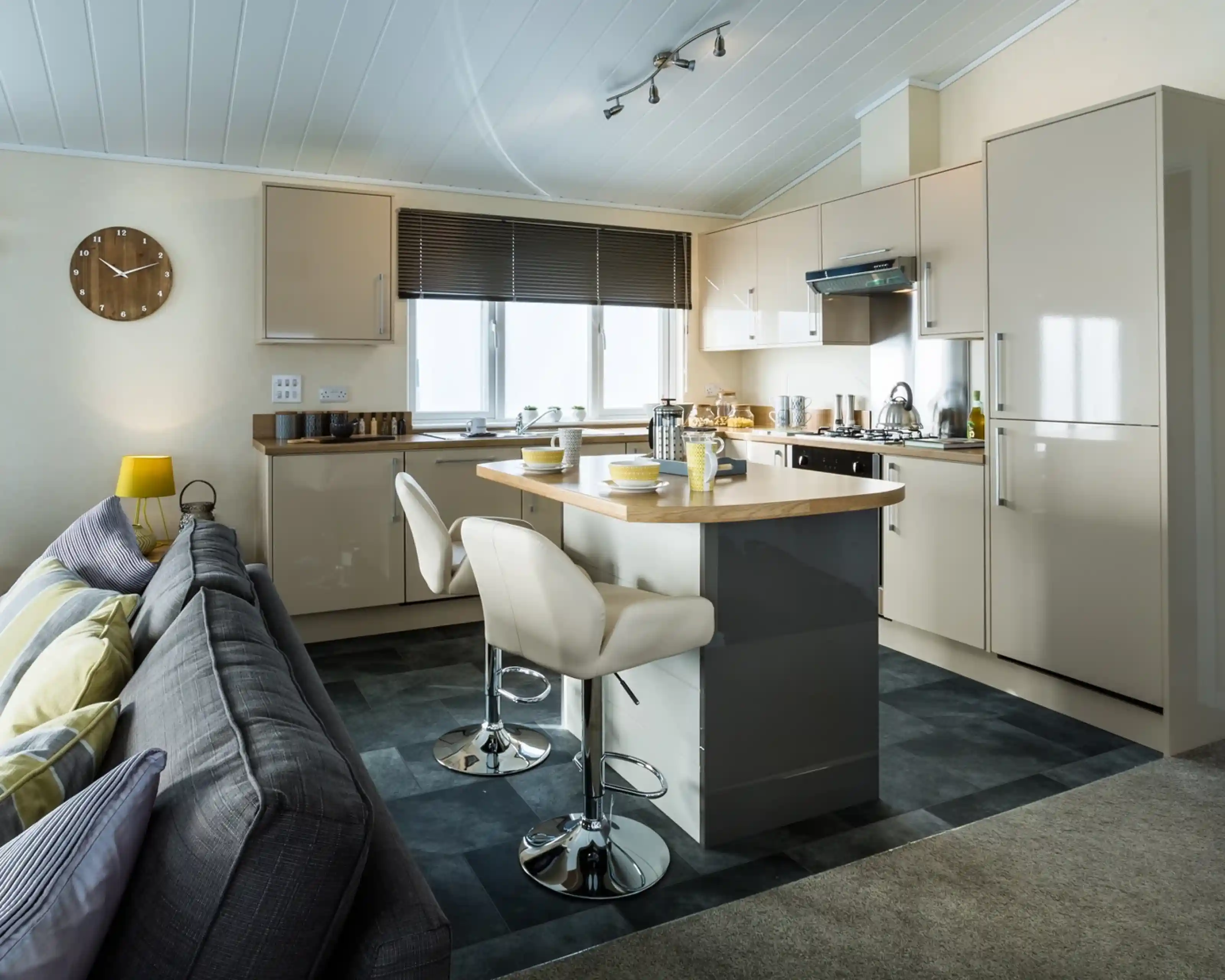 A bright modern kitchen with glossy cream cabinets, a stainless steel oven and hood, and a wooden breakfast bar with two cream stools. The bar is set with mugs and bowls, while a sofa with striped cushions is visible in the foreground. A yellow lamp and wooden wall clock add warmth to the neutral décor, and large windows let in natural light.