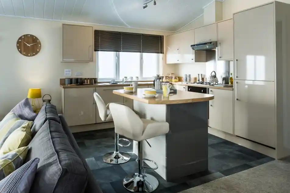 A bright modern kitchen with glossy cream cabinets, a stainless steel oven and hood, and a wooden breakfast bar with two cream stools. The bar is set with mugs and bowls, while a sofa with striped cushions is visible in the foreground. A yellow lamp and wooden wall clock add warmth to the neutral décor, and large windows let in natural light.