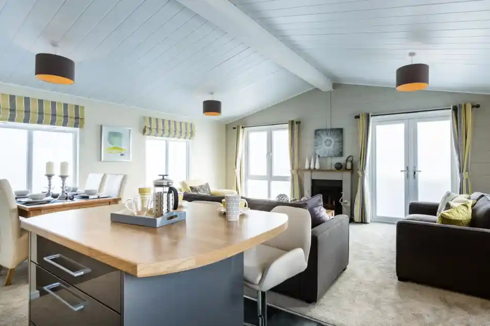 A bright and modern open-plan lounge, kitchen, diner with a wooden kitchen island with breakfast bar in the foreground. A carpeted lounge with two grey sofas, a fireplace and a dining table sit in the background, together with full length windows and doors allowing plenty of natural light. The room features yellow and grey accents adding character to the space.