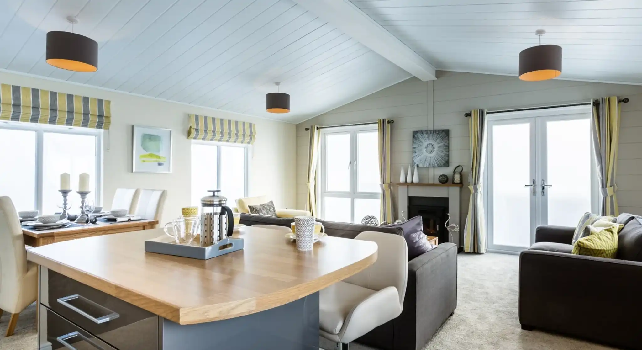A bright and modern open-plan lounge, kitchen, diner with a wooden kitchen island with breakfast bar in the foreground. A carpeted lounge with two grey sofas, a fireplace and a dining table sit in the background, together with full length windows and doors allowing plenty of natural light. The room features yellow and grey accents adding character to the space.