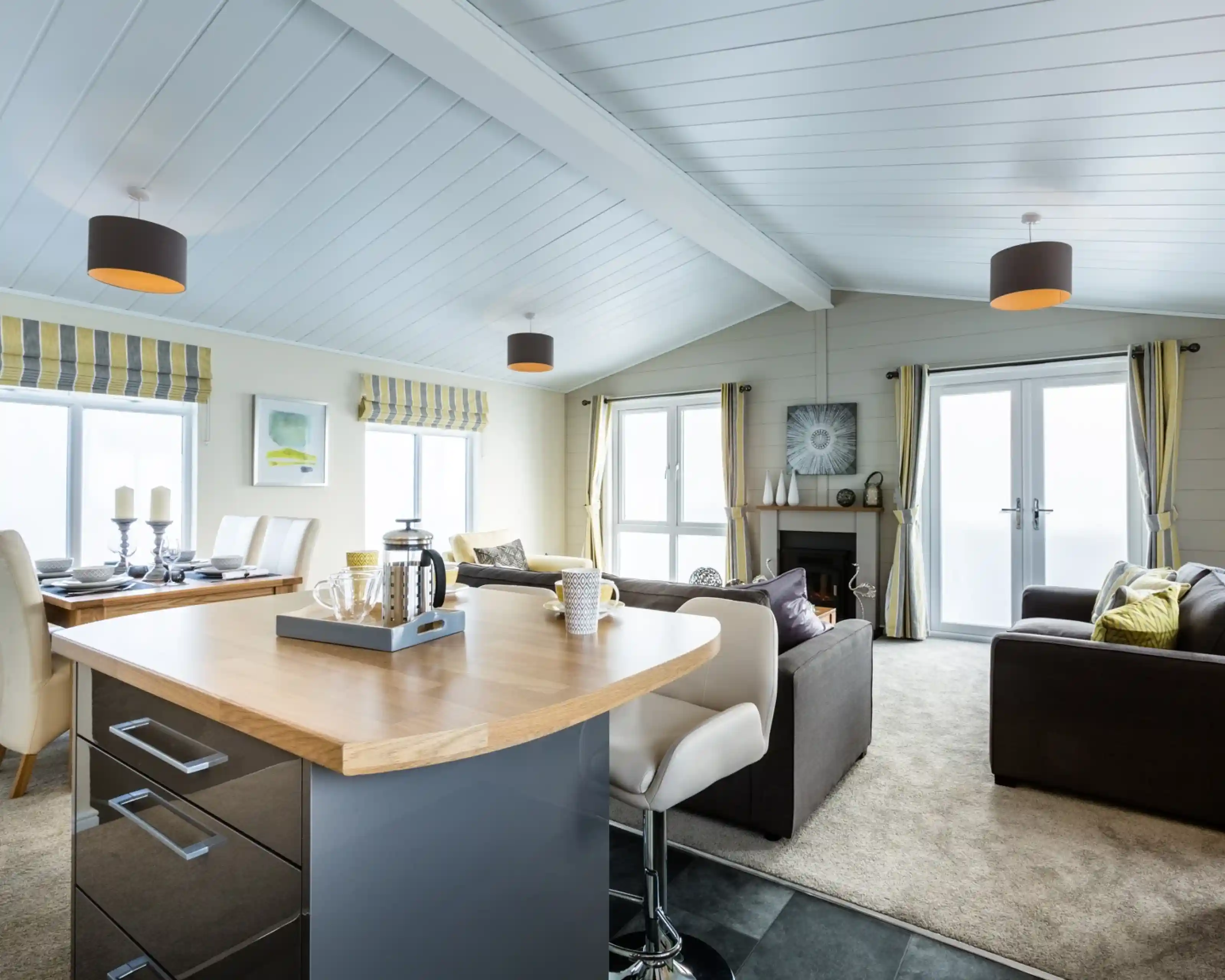 A bright and modern open-plan lounge, kitchen, diner with a wooden kitchen island with breakfast bar in the foreground. A carpeted lounge with two grey sofas, a fireplace and a dining table sit in the background, together with full length windows and doors allowing plenty of natural light. The room features yellow and grey accents adding character to the space.