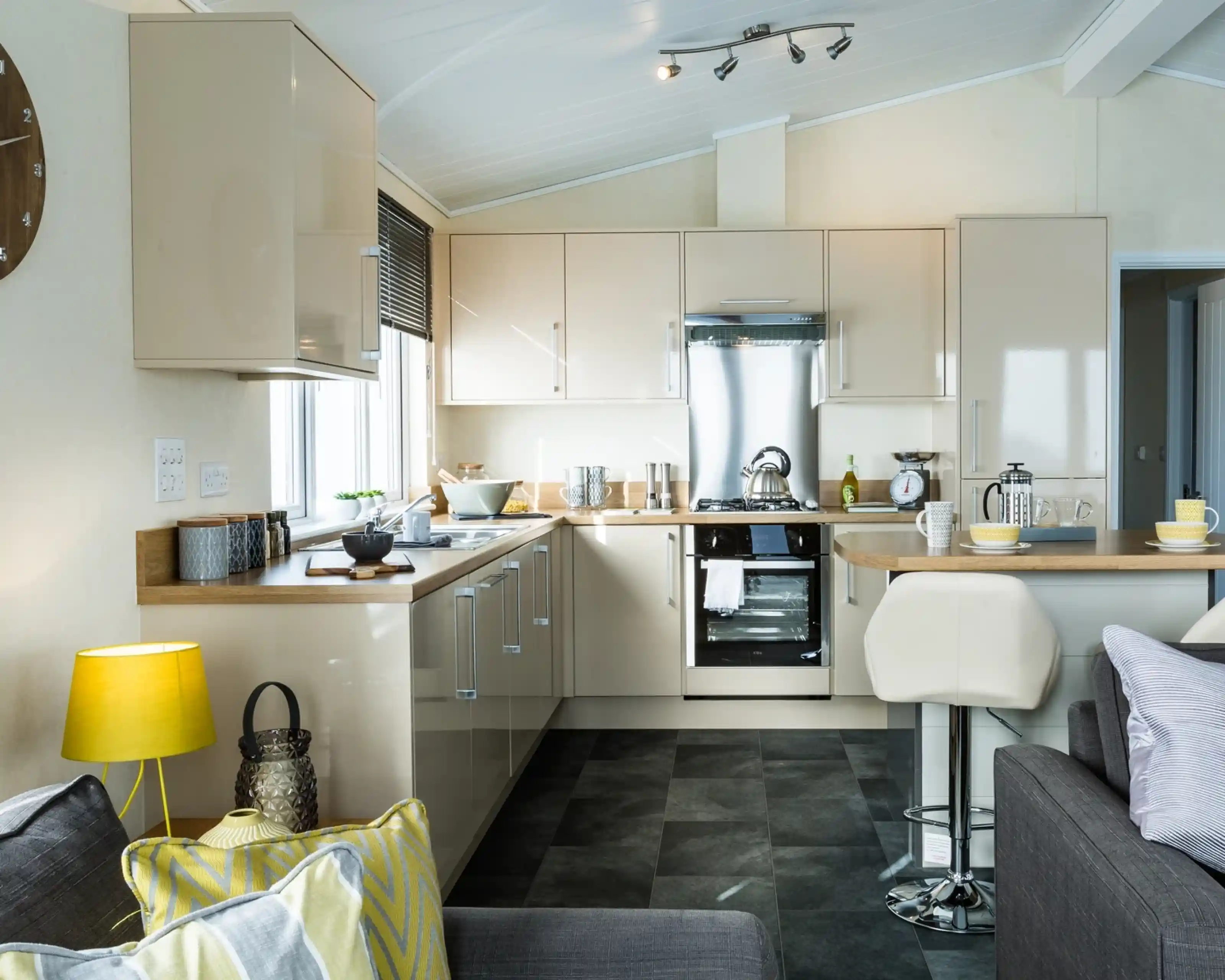 A modern kitchen with glossy cream cabinets, a stainless steel oven, and a matching extractor hood. Wooden countertops run along two sides, with small appliances and utensils neatly arranged. A breakfast bar with two white stools extends into the room, set with cups and bowls. The kitchen is well lit with natural light from a window and ceiling spotlights, and part of a cosy living area with a sofa, cushions, and a yellow lamp is visible in the foreground.