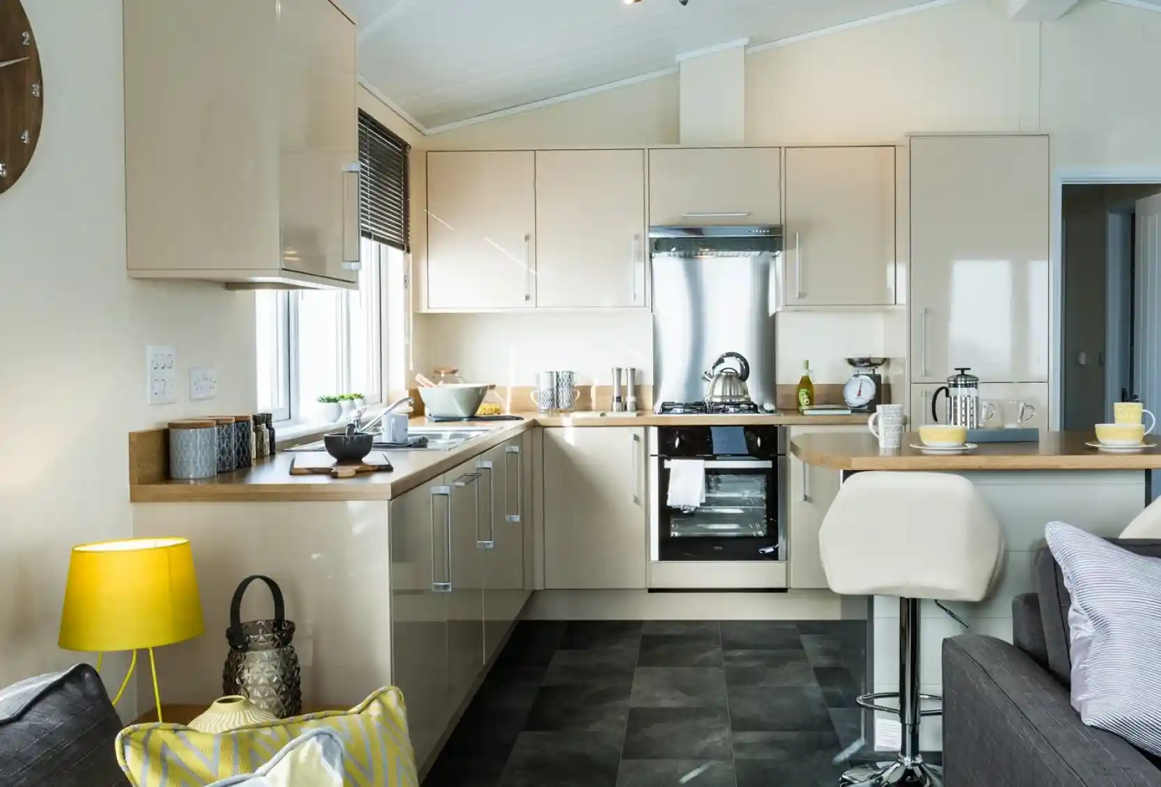 A modern kitchen with glossy cream cabinets, a stainless steel oven, and a matching extractor hood. Wooden countertops run along two sides, with small appliances and utensils neatly arranged. A breakfast bar with two white stools extends into the room, set with cups and bowls. The kitchen is well lit with natural light from a window and ceiling spotlights, and part of a cosy living area with a sofa, cushions, and a yellow lamp is visible in the foreground.