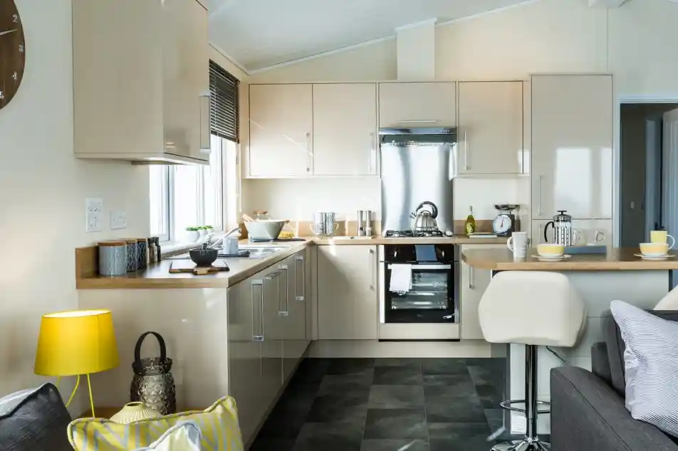 A modern kitchen with glossy cream cabinets, a stainless steel oven, and a matching extractor hood. Wooden countertops run along two sides, with small appliances and utensils neatly arranged. A breakfast bar with two white stools extends into the room, set with cups and bowls. The kitchen is well lit with natural light from a window and ceiling spotlights, and part of a cosy living area with a sofa, cushions, and a yellow lamp is visible in the foreground.
