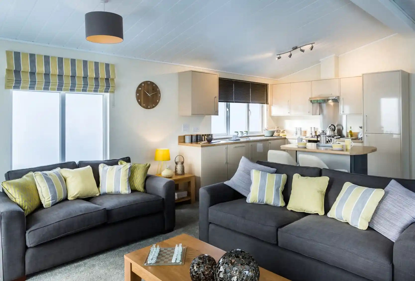 A modern open-plan living room and kitchen. The lounge features two dark grey sofas with striped and yellow cushions, a wooden coffee table with decorative ornaments, and a chessboard. Behind the sofas, the kitchen has cream cabinets, a stainless steel oven, and a breakfast bar with stools. A striped Roman blind covers one window, and a grey ceiling light and yellow lamp add warmth to the space.
