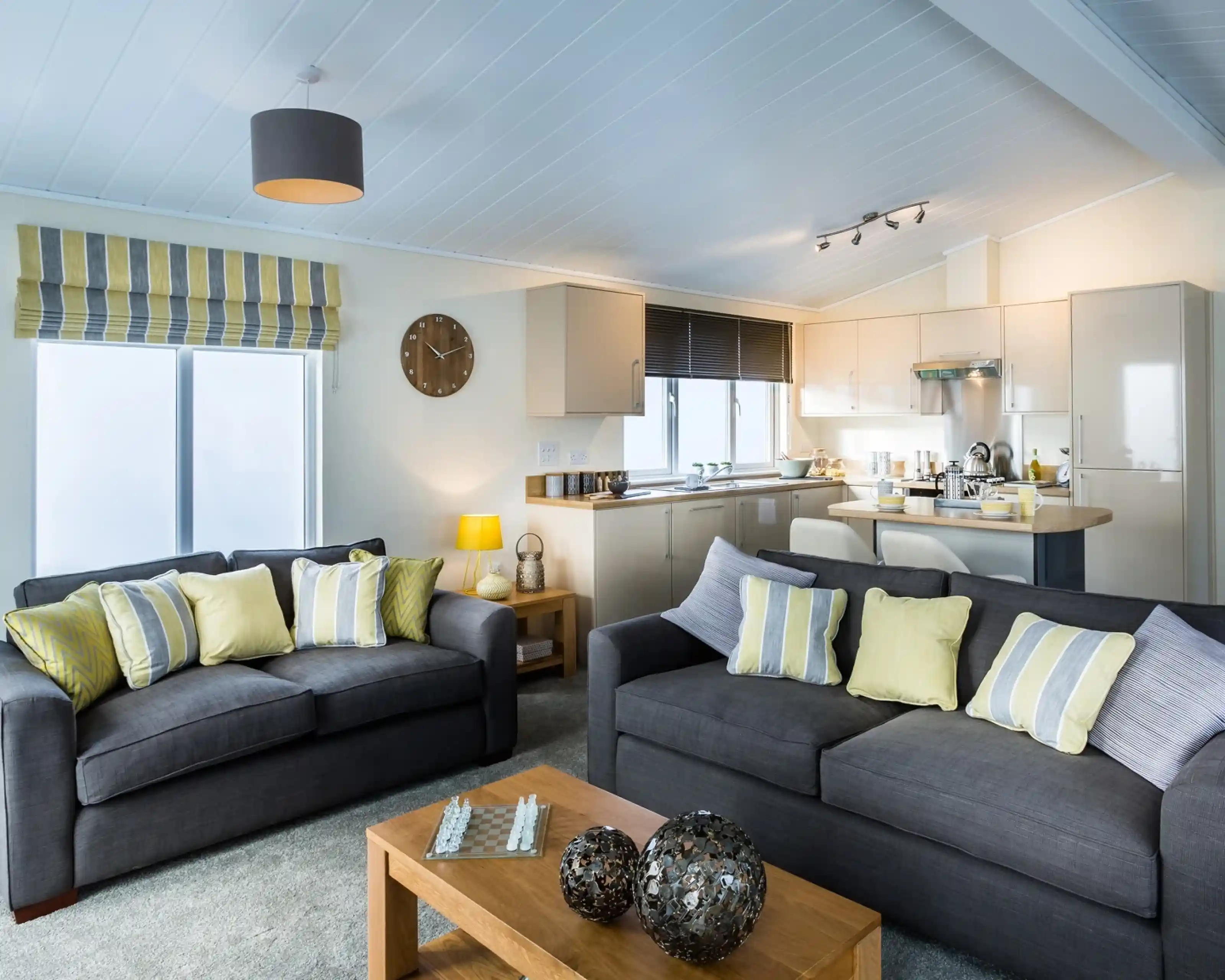 A modern open-plan living room and kitchen. The lounge features two dark grey sofas with striped and yellow cushions, a wooden coffee table with decorative ornaments, and a chessboard. Behind the sofas, the kitchen has cream cabinets, a stainless steel oven, and a breakfast bar with stools. A striped Roman blind covers one window, and a grey ceiling light and yellow lamp add warmth to the space.