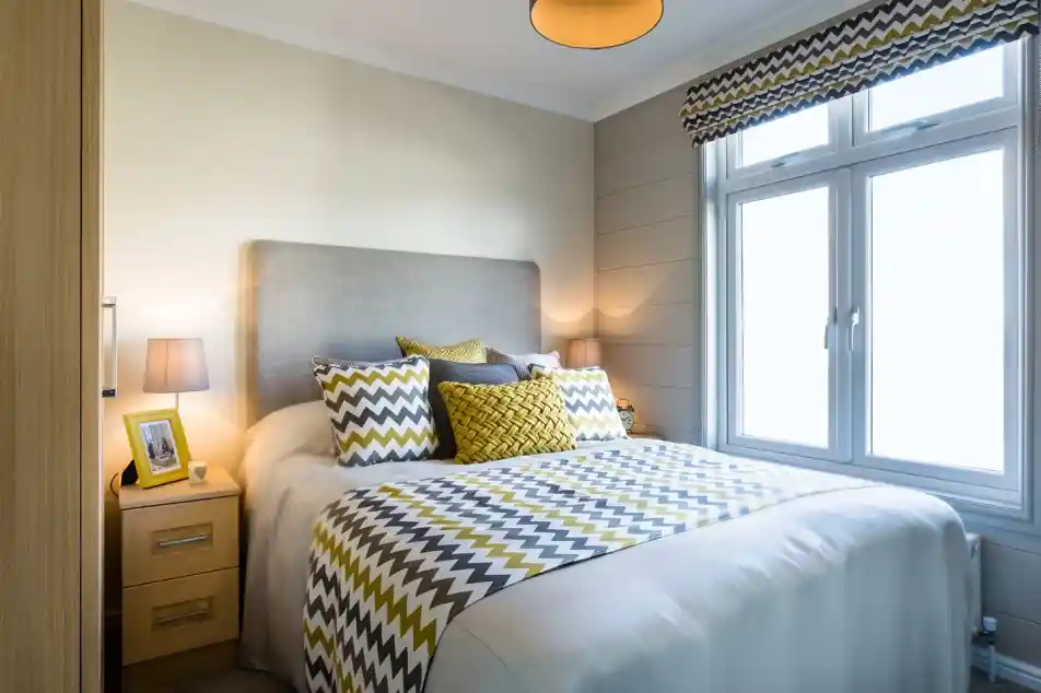 A light and airy bedroom with a double bed with crisp white sheets, and yellow and grey patterned cushions and bedspread. A yellow photoframe, grey ceiling pendant and patterned blind complements the stylish room. there is also two bedside tables with matching lamps and a wardrobe.