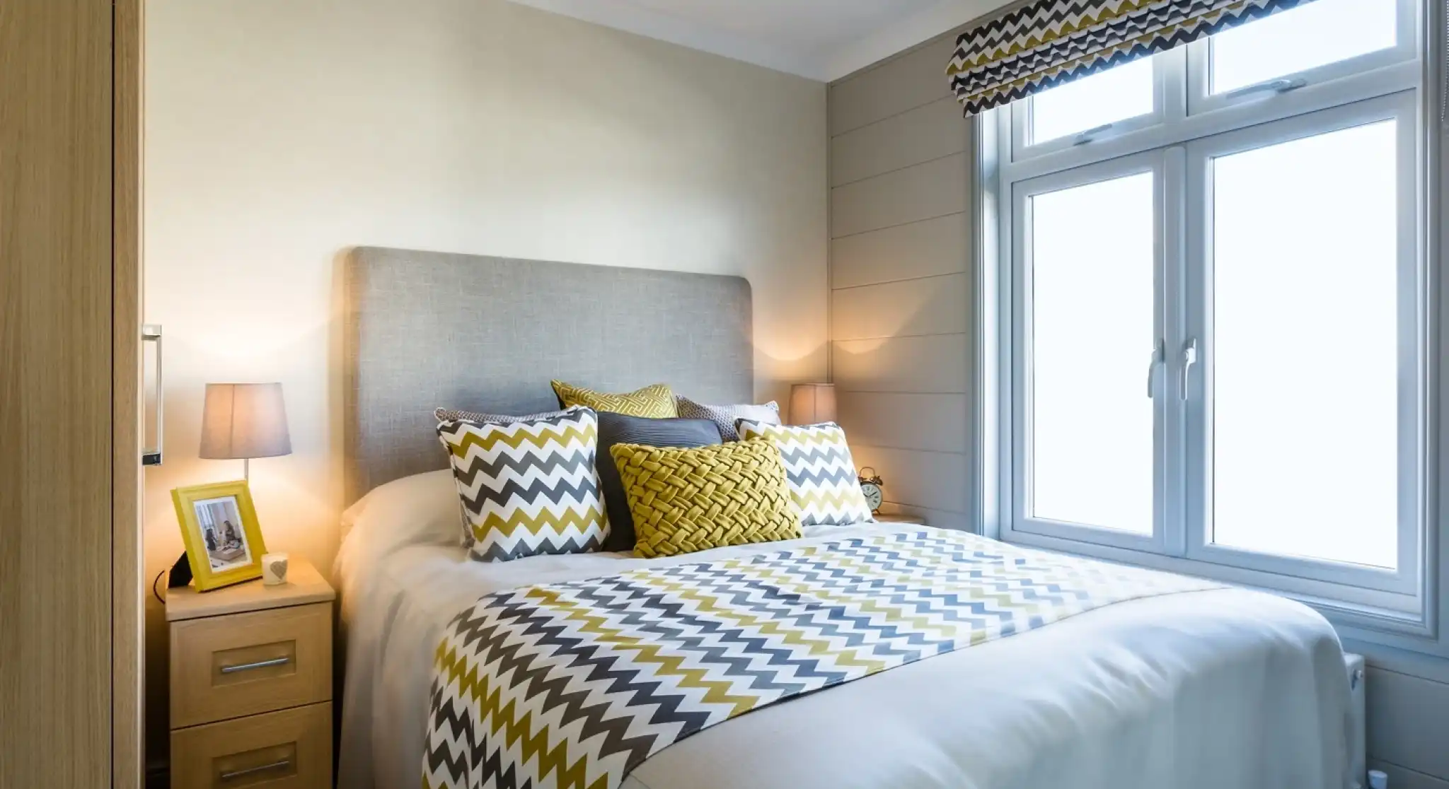 A light and airy bedroom with a double bed with crisp white sheets, and yellow and grey patterned cushions and bedspread. A yellow photoframe, grey ceiling pendant and patterned blind complements the stylish room. there is also two bedside tables with matching lamps and a wardrobe.