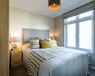 A light and airy bedroom with a double bed with crisp white sheets, and yellow and grey patterned cushions and bedspread. A yellow photoframe, grey ceiling pendant and patterned blind complements the stylish room. there is also two bedside tables with matching lamps and a wardrobe.