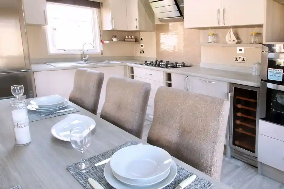 A modern kitchen featuring light-colored cabinets, a stainless steel refrigerator, and a gas stove. A dining table is set with plates and wine glasses, accompanied by upholstered chairs. The space is bright with natural light.