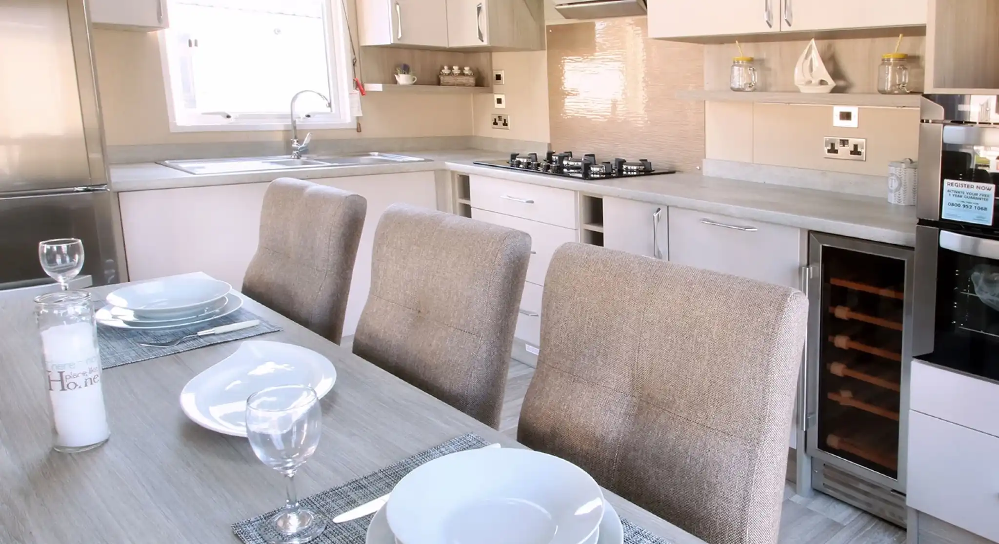 A modern kitchen featuring light-colored cabinets, a stainless steel refrigerator, and a gas stove. A dining table is set with plates and wine glasses, accompanied by upholstered chairs. The space is bright with natural light.