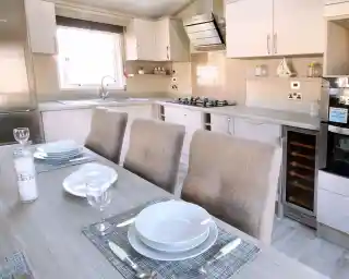A modern kitchen featuring light-colored cabinets, a stainless steel refrigerator, and a gas stove. A dining table is set with plates and wine glasses, accompanied by upholstered chairs. The space is bright with natural light.