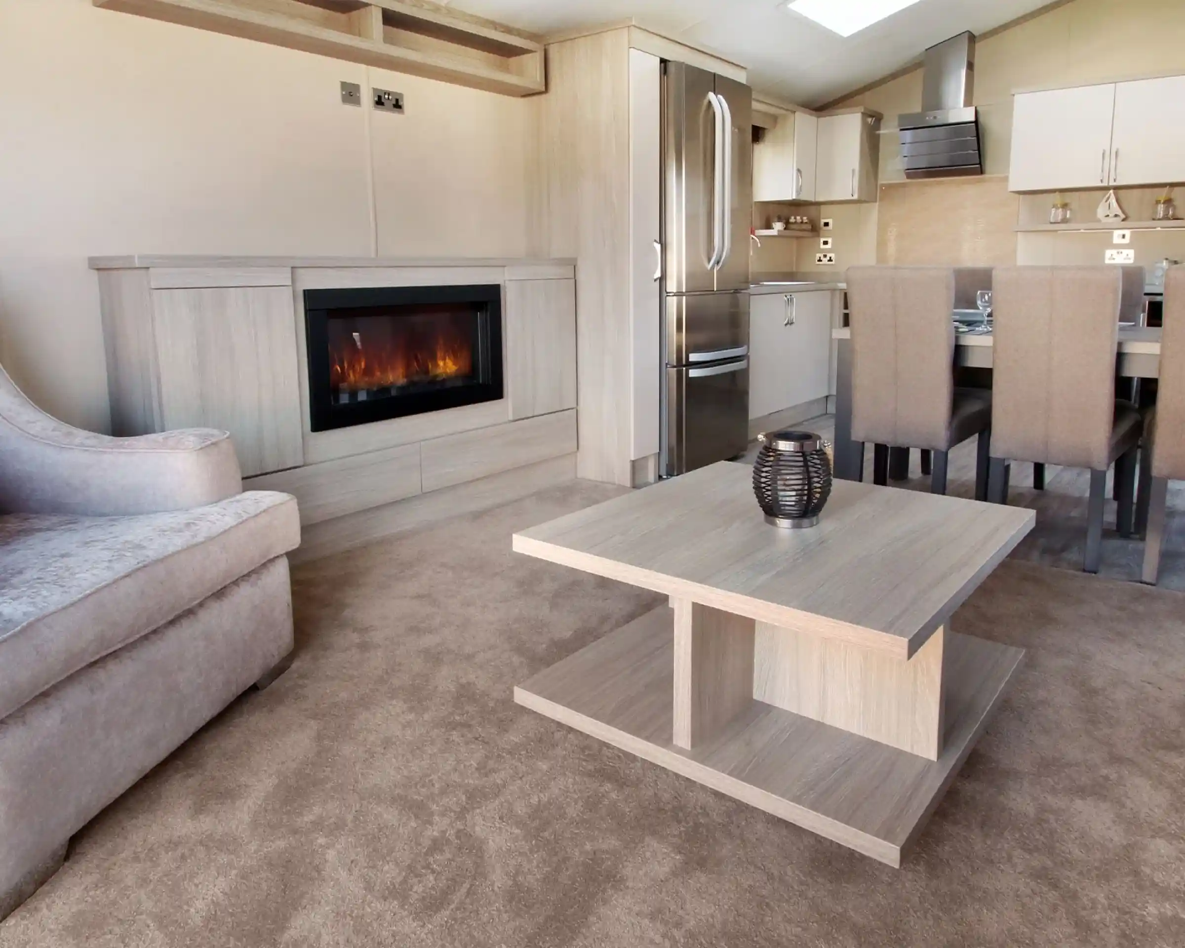 A modern living space featuring a plush gray sofa, a sleek wooden coffee table, and a stylish electric fireplace. The kitchen area is visible in the background, with a stainless steel refrigerator and dining table set for six. The flooring is covered in light beige carpet, enhancing the cozy atmosphere.