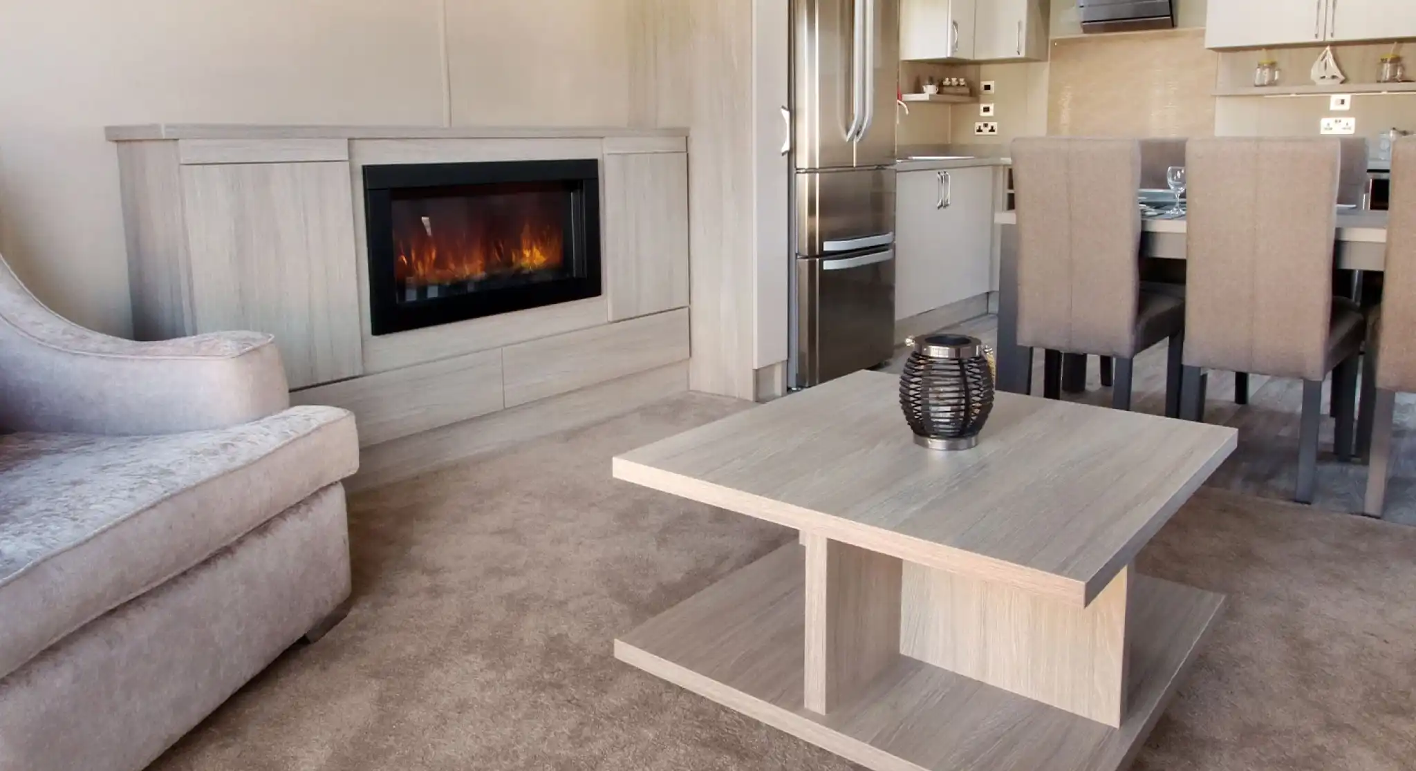 A modern living space featuring a plush gray sofa, a sleek wooden coffee table, and a stylish electric fireplace. The kitchen area is visible in the background, with a stainless steel refrigerator and dining table set for six. The flooring is covered in light beige carpet, enhancing the cozy atmosphere.
