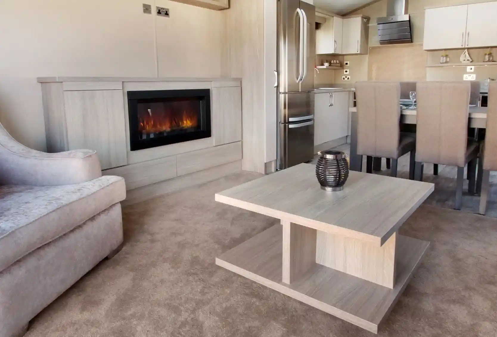 A modern living space featuring a plush gray sofa, a sleek wooden coffee table, and a stylish electric fireplace. The kitchen area is visible in the background, with a stainless steel refrigerator and dining table set for six. The flooring is covered in light beige carpet, enhancing the cozy atmosphere.