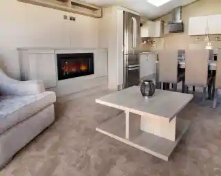 A modern living space featuring a plush gray sofa, a sleek wooden coffee table, and a stylish electric fireplace. The kitchen area is visible in the background, with a stainless steel refrigerator and dining table set for six. The flooring is covered in light beige carpet, enhancing the cozy atmosphere.