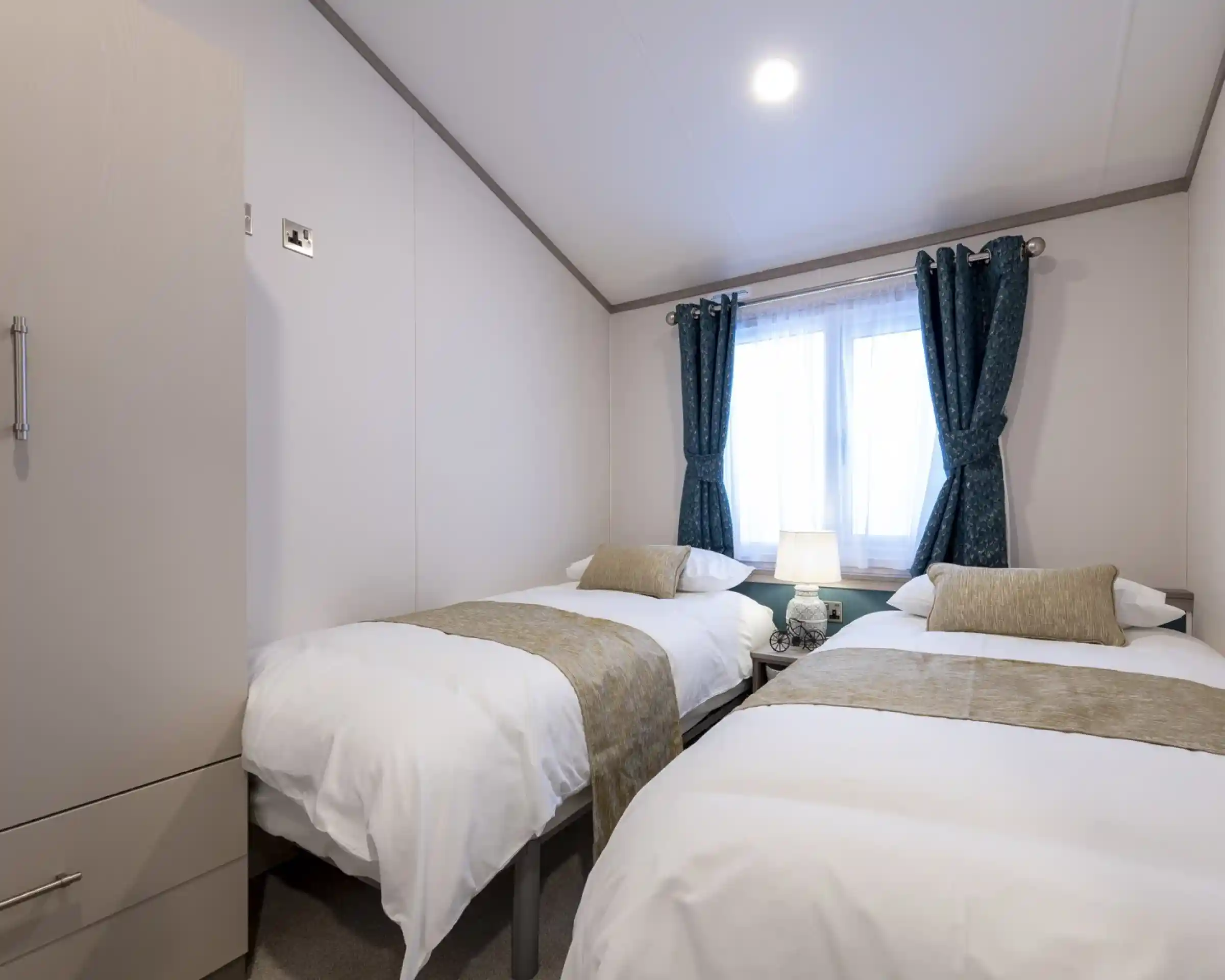 A light, bright twin bedroom with twin single beds with crisp white linen and gold cushions and throws, together with a bedside table, a wardrobe and blue colours curtains at the window.