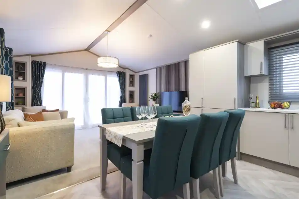 A large and airy room featuring a dining table with teal coloured upholstered dining chairs and wooden table on a herringbone style floor with kitchen cabinets to one side. In the background is a lounge with neutral coloured sofas and carpets, together with a TV and patio doors with side screens.