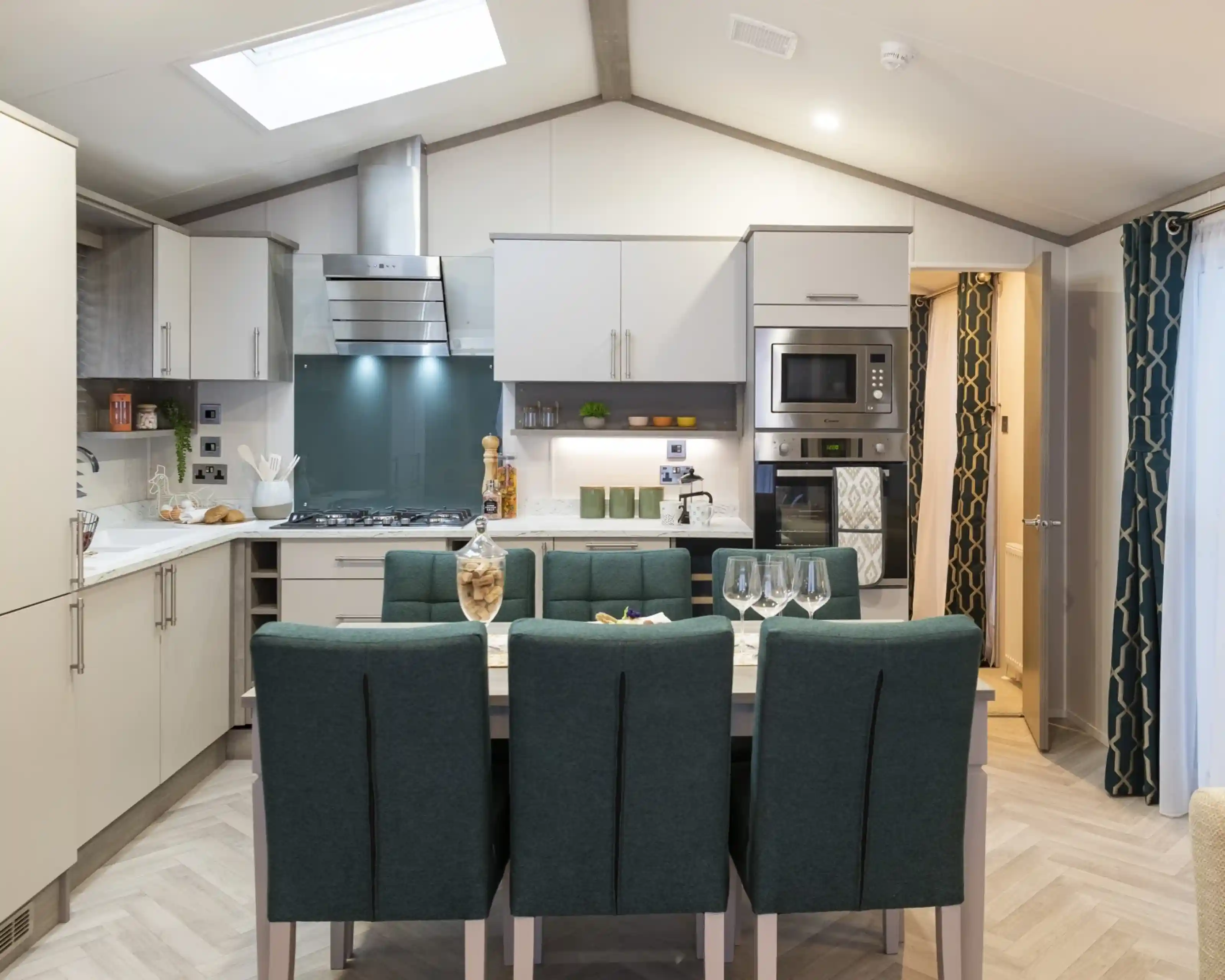 A bright kitchen with modern neutral coloured cabinets and built-in appliances. There is dining table with teal coloured upholstered dining chairs. The room also has a Velux window allowing natural light in, together with herringbone wooden style flooring.
