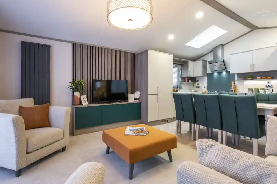 A large open-plan living space with neutral sofas and carpet to the lounge area, together with a rust coloured foot stool/coffee table, and feature wooden slatted wall with built-in teal painted TV cabinet with TV. To one side is the dining and kitchen area with matching neutral coloured cabinets and teal colour dining chairs.