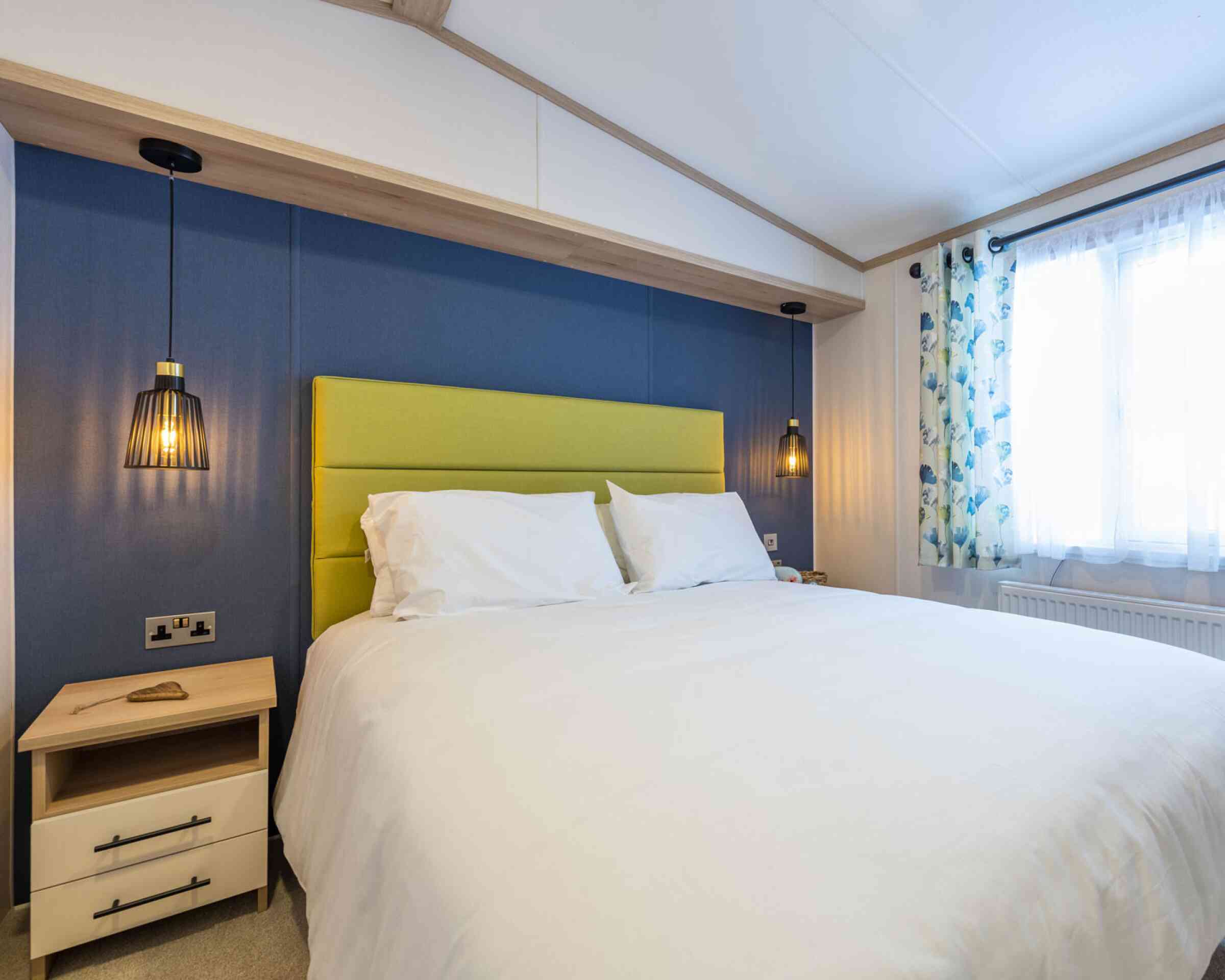 A stylish bedroom showcasing a double bed with white bed linen set against vibrant blue walls and a lime green upholstered headboard. Two bedside tables flank the bed with matching pendant ceiling beside lights.