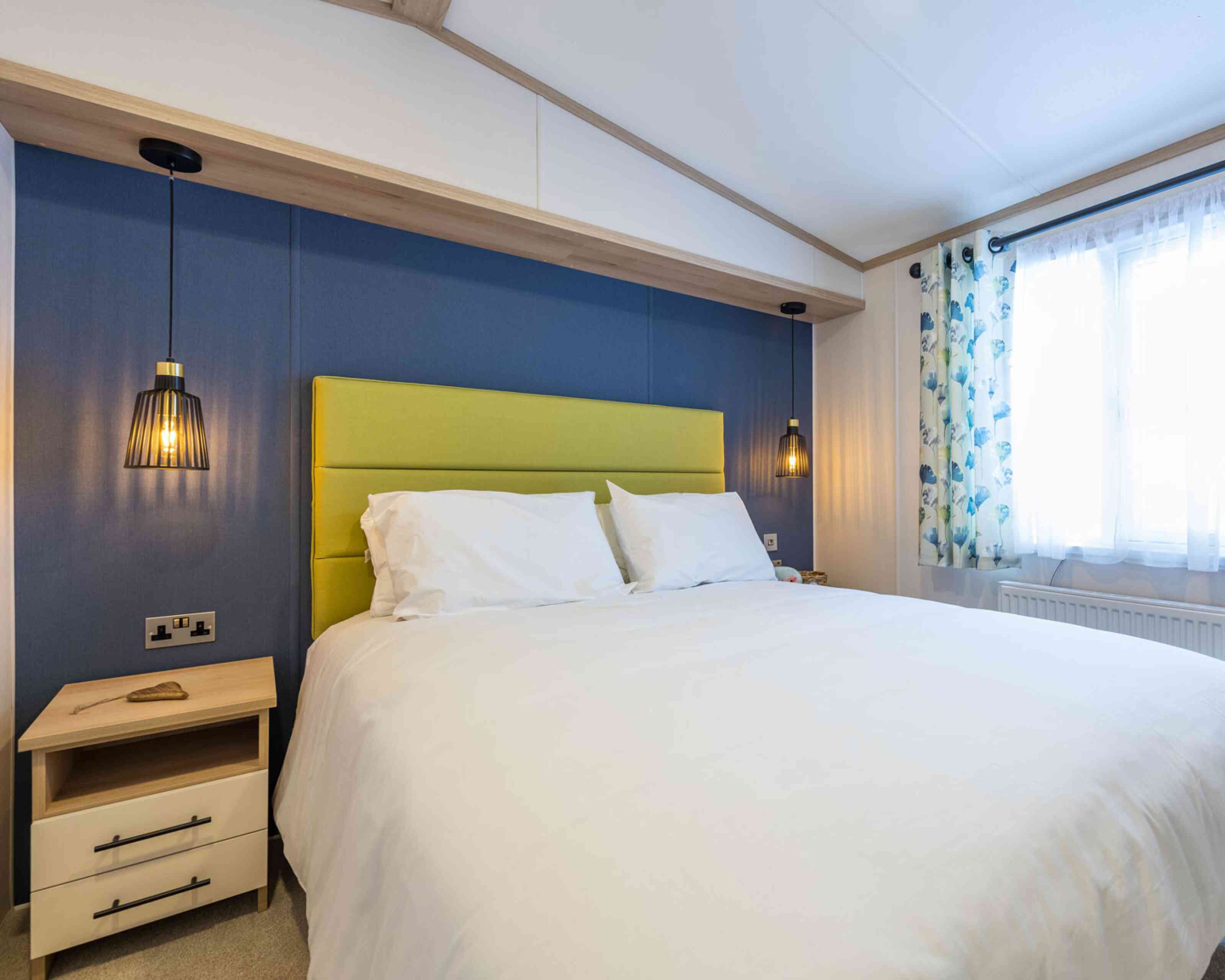 A stylish bedroom showcasing a double bed with white bed linen set against vibrant blue walls and a lime green upholstered headboard. Two bedside tables flank the bed with matching pendant ceiling beside lights.