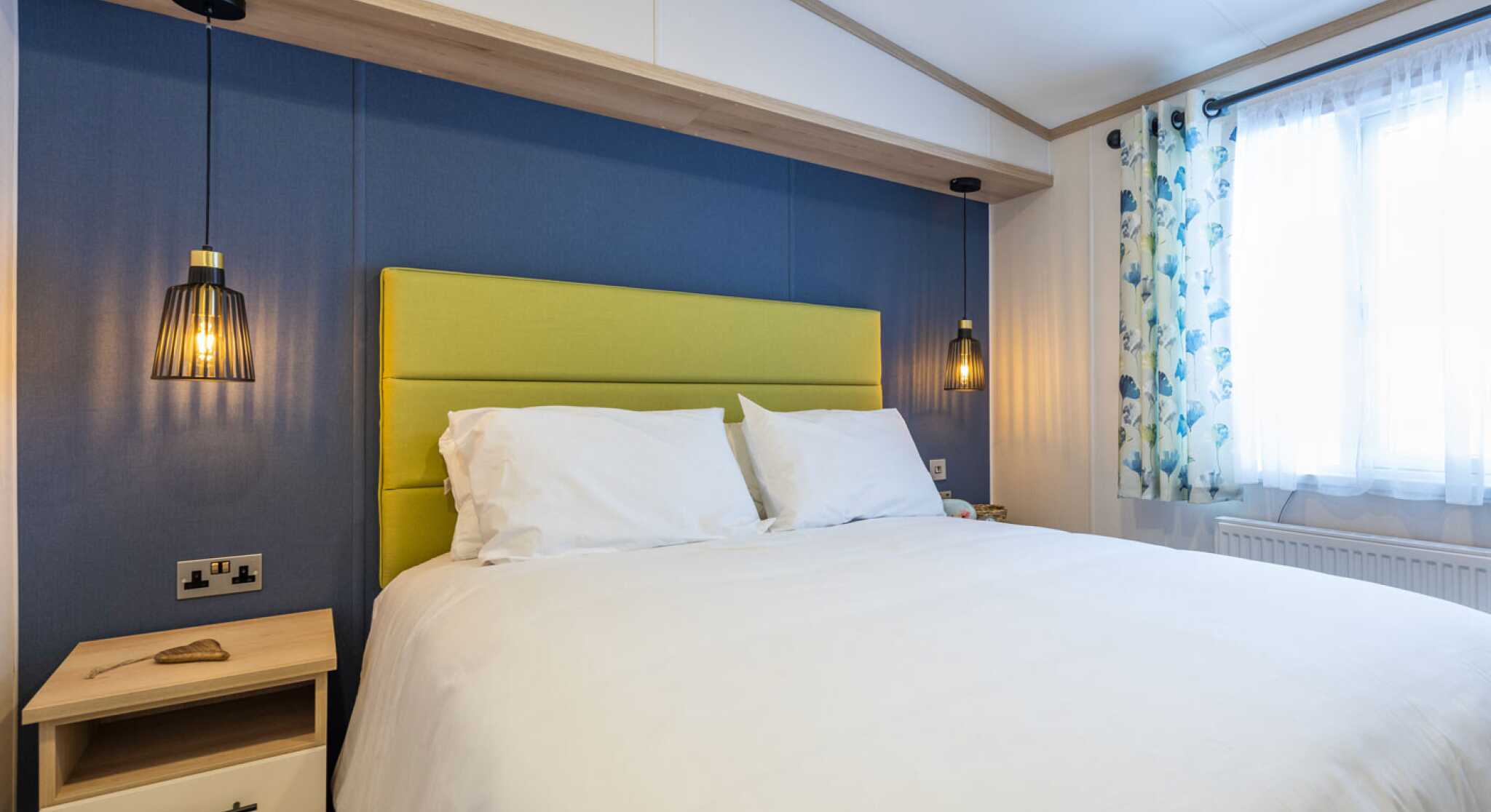 A stylish bedroom showcasing a double bed with white bed linen set against vibrant blue walls and a lime green upholstered headboard. Two bedside tables flank the bed with matching pendant ceiling beside lights.