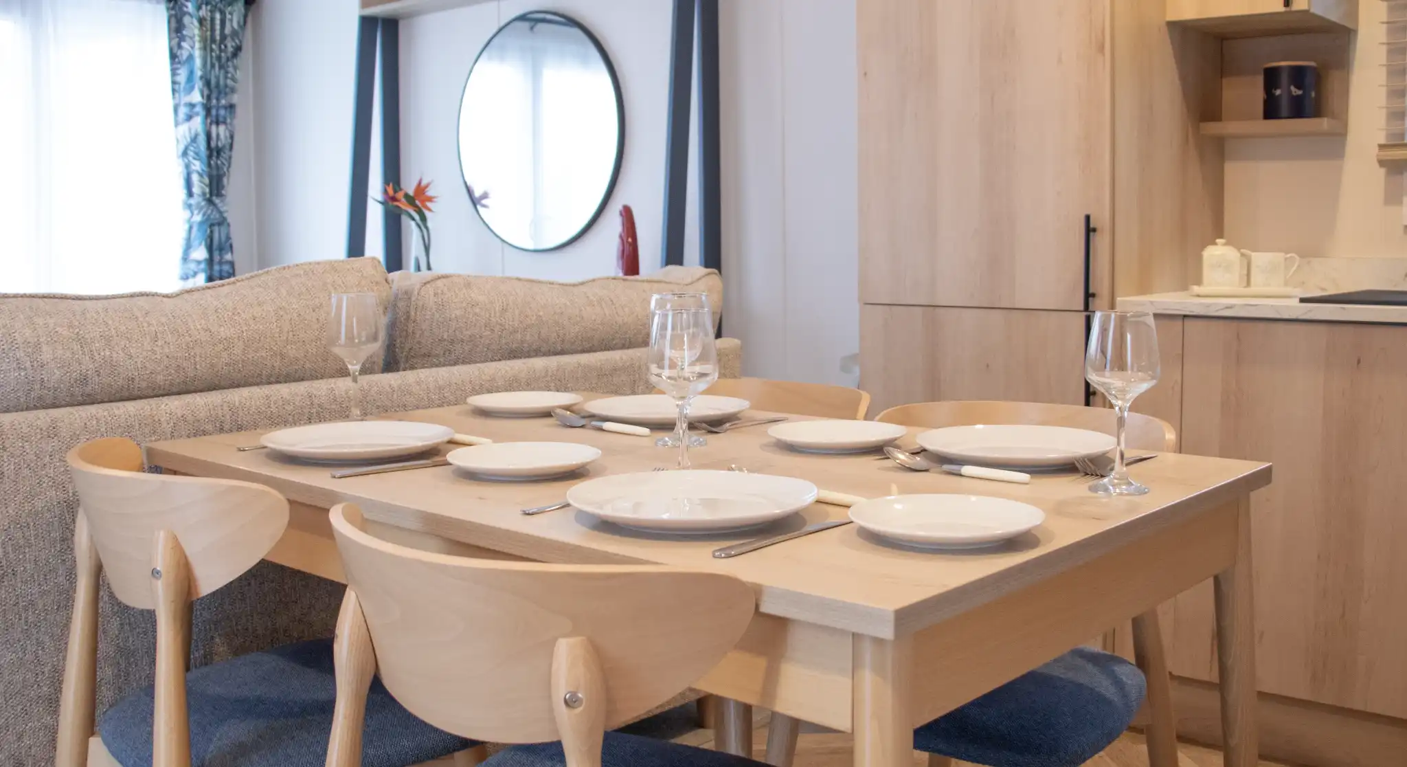 A modern dining table set for six, featuring white plates, wine glasses, and cutlery. The table is surrounded by light wood chairs with blue cushions, beside a cozy sofa. A round mirror and wooden cabinets are visible in the background.