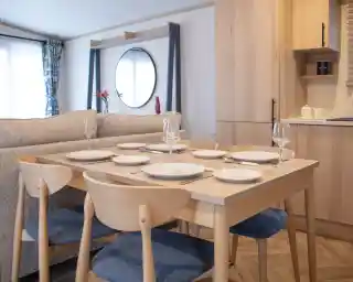 A modern dining table set for six, featuring white plates, wine glasses, and cutlery. The table is surrounded by light wood chairs with blue cushions, beside a cozy sofa. A round mirror and wooden cabinets are visible in the background.