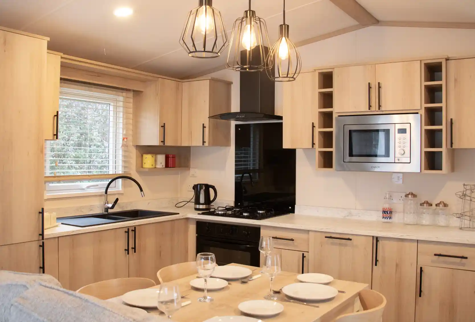 A modern kitchen with wooden cabinetry, featuring a black sink and stove, a microwave, and under-cabinet lighting. A dining table with set dinnerware is in the foreground, with three stylish hanging pendant lights above. Bright natural light streams in through a window.