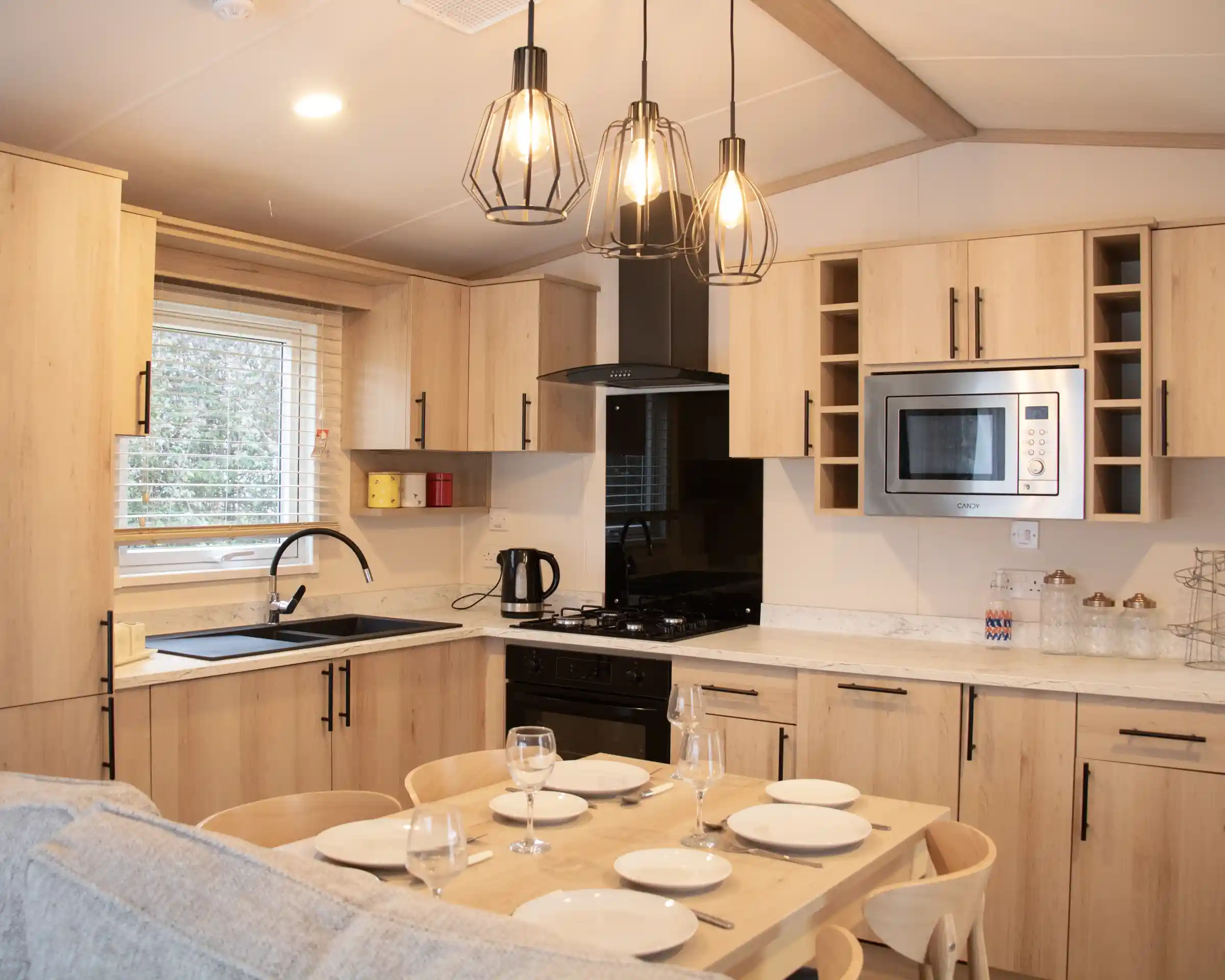 A modern kitchen with wooden cabinetry, featuring a black sink and stove, a microwave, and under-cabinet lighting. A dining table with set dinnerware is in the foreground, with three stylish hanging pendant lights above. Bright natural light streams in through a window.