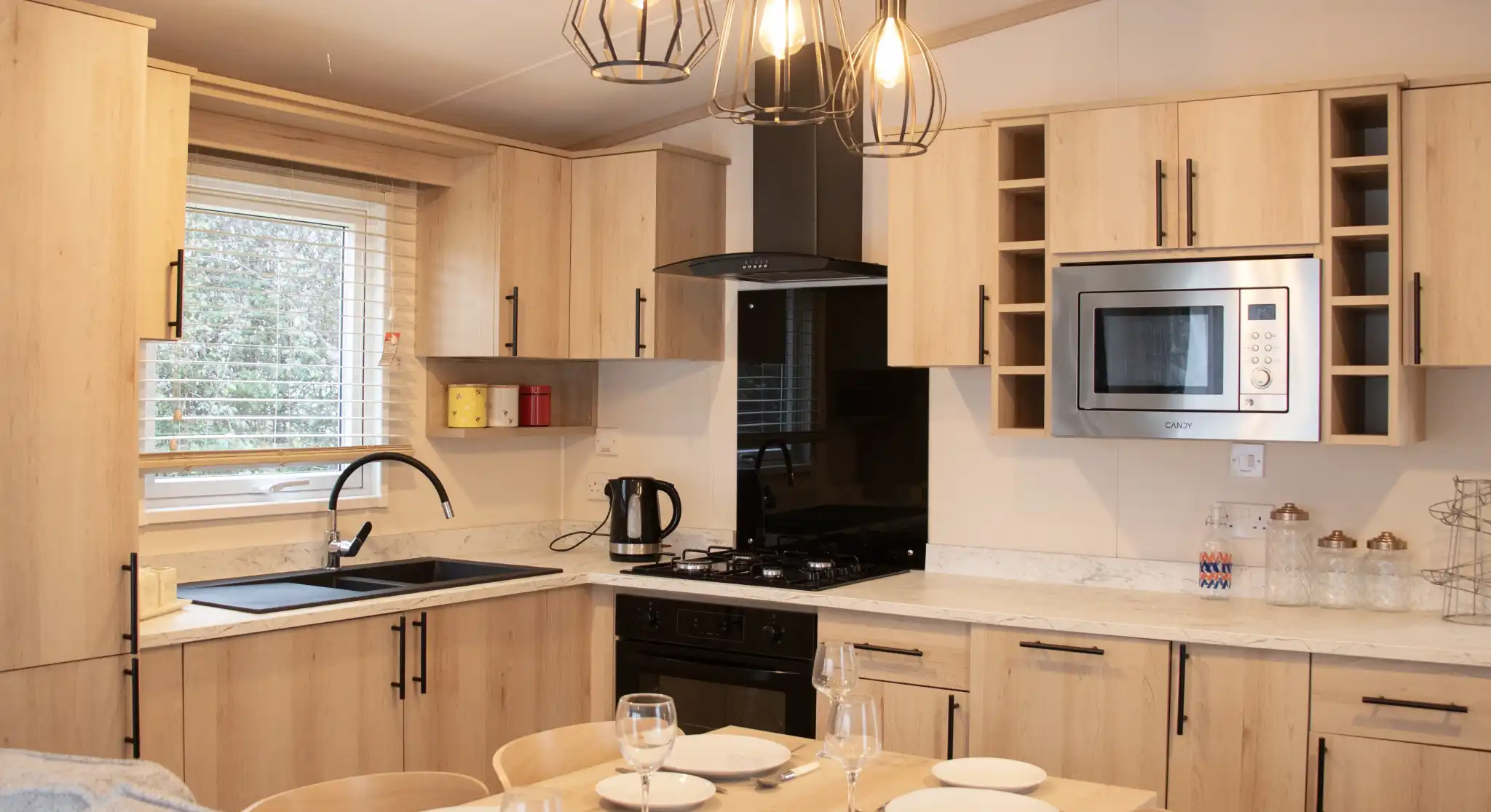 A modern kitchen with wooden cabinetry, featuring a black sink and stove, a microwave, and under-cabinet lighting. A dining table with set dinnerware is in the foreground, with three stylish hanging pendant lights above. Bright natural light streams in through a window.