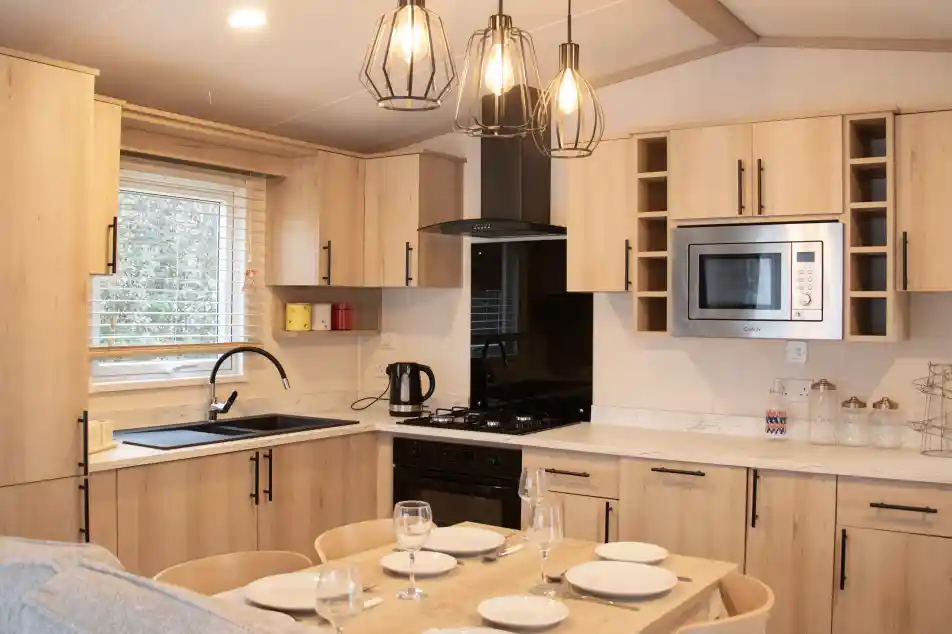 A modern kitchen with wooden cabinetry, featuring a black sink and stove, a microwave, and under-cabinet lighting. A dining table with set dinnerware is in the foreground, with three stylish hanging pendant lights above. Bright natural light streams in through a window.