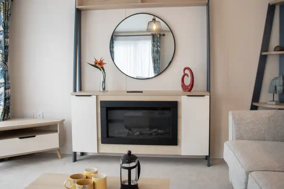 Modern living room featuring a stylish wall-mounted cabinet with a circular mirror above. An electric fireplace is centered within the cabinet, complemented by decorative items, including a vase with a flower and a sculptural piece. A light-colored sofa and a coffee table with yellow mugs and a lantern enhance the cozy atmosphere. Natural light floods in through a window with patterned curtains.