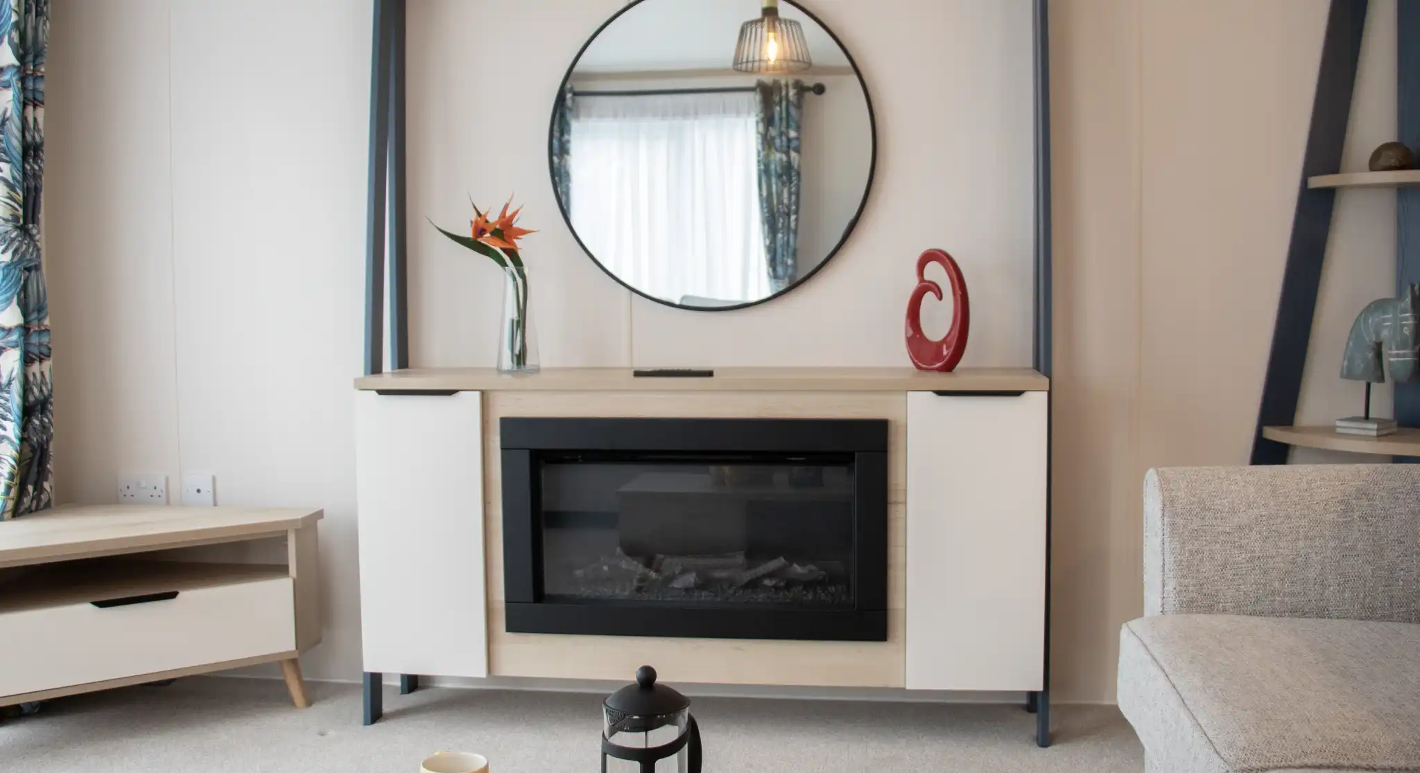 Modern living room featuring a stylish wall-mounted cabinet with a circular mirror above. An electric fireplace is centered within the cabinet, complemented by decorative items, including a vase with a flower and a sculptural piece. A light-colored sofa and a coffee table with yellow mugs and a lantern enhance the cozy atmosphere. Natural light floods in through a window with patterned curtains.
