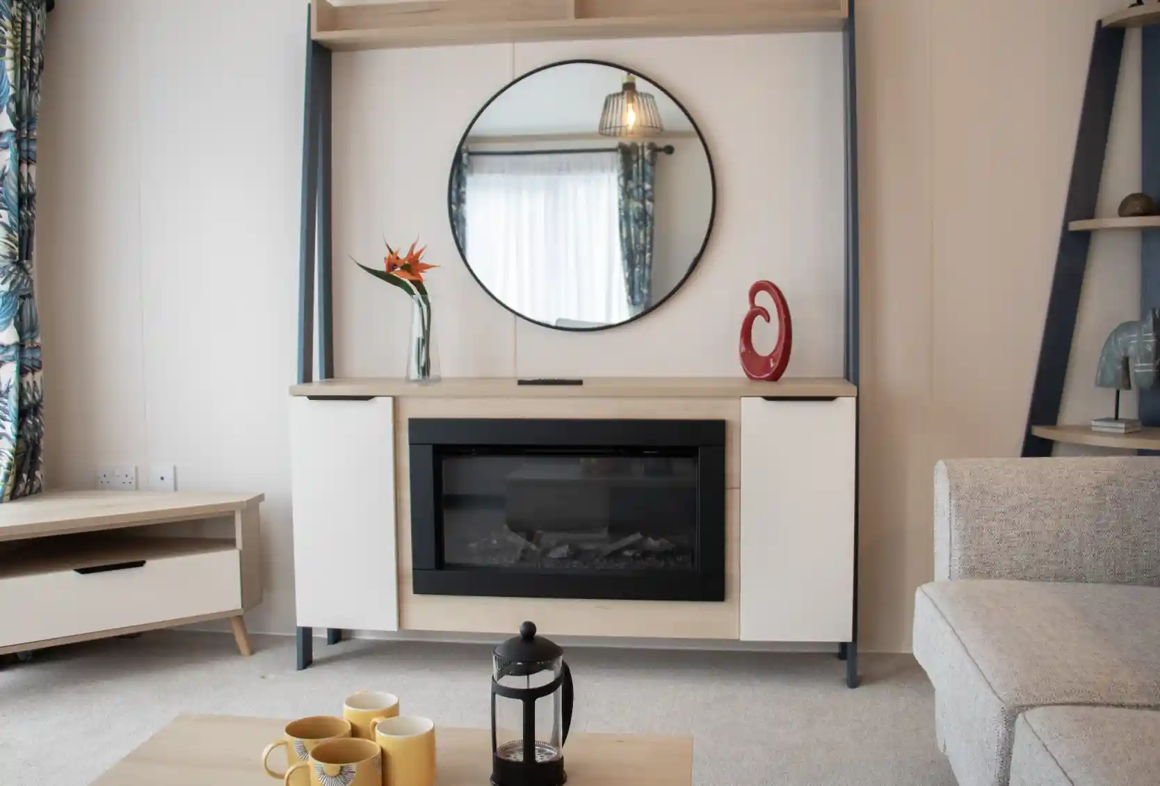Modern living room featuring a stylish wall-mounted cabinet with a circular mirror above. An electric fireplace is centered within the cabinet, complemented by decorative items, including a vase with a flower and a sculptural piece. A light-colored sofa and a coffee table with yellow mugs and a lantern enhance the cozy atmosphere. Natural light floods in through a window with patterned curtains.