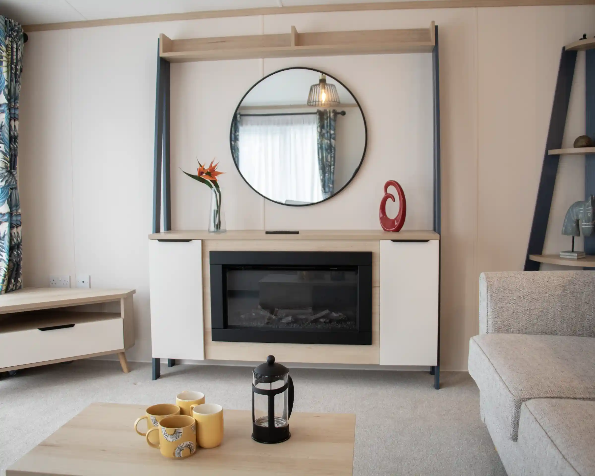 Modern living room featuring a stylish wall-mounted cabinet with a circular mirror above. An electric fireplace is centered within the cabinet, complemented by decorative items, including a vase with a flower and a sculptural piece. A light-colored sofa and a coffee table with yellow mugs and a lantern enhance the cozy atmosphere. Natural light floods in through a window with patterned curtains.