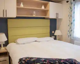 A cozy bedroom featuring a green upholstered headboard, white bedding, and decorative pillows. Shelves above the bed hold a small vase and a lantern, while a lamp and a patterned blanket are on the bedside table. Natural light filters through a window with floral curtains.