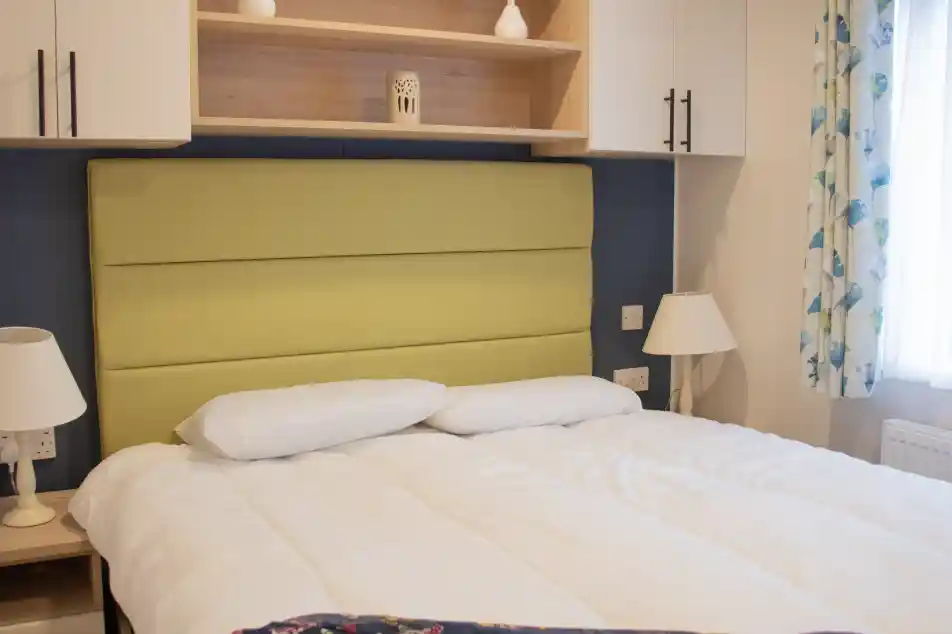 A cozy bedroom featuring a green upholstered headboard, white bedding, and decorative pillows. Shelves above the bed hold a small vase and a lantern, while a lamp and a patterned blanket are on the bedside table. Natural light filters through a window with floral curtains.