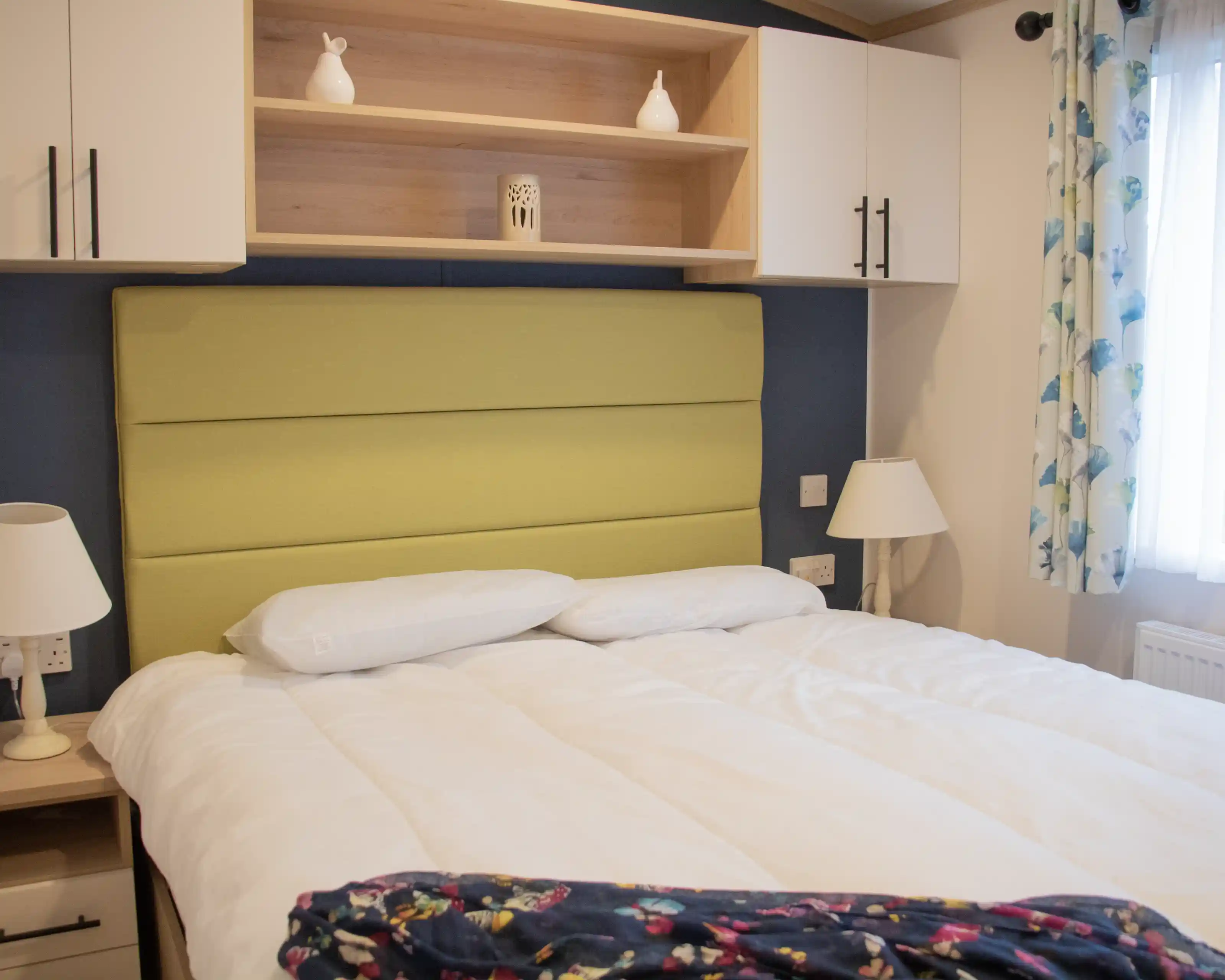 A cozy bedroom featuring a green upholstered headboard, white bedding, and decorative pillows. Shelves above the bed hold a small vase and a lantern, while a lamp and a patterned blanket are on the bedside table. Natural light filters through a window with floral curtains.