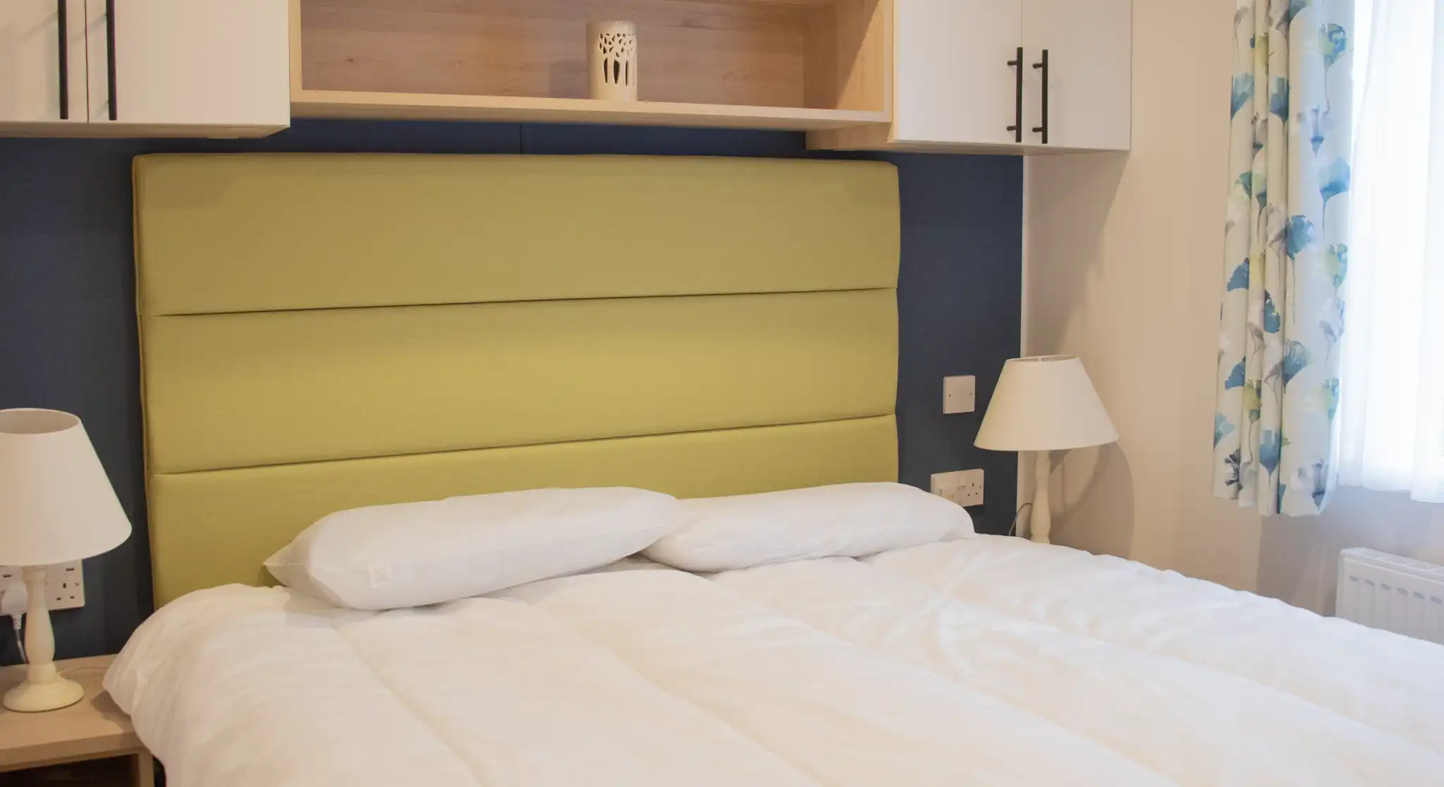 A cozy bedroom featuring a green upholstered headboard, white bedding, and decorative pillows. Shelves above the bed hold a small vase and a lantern, while a lamp and a patterned blanket are on the bedside table. Natural light filters through a window with floral curtains.