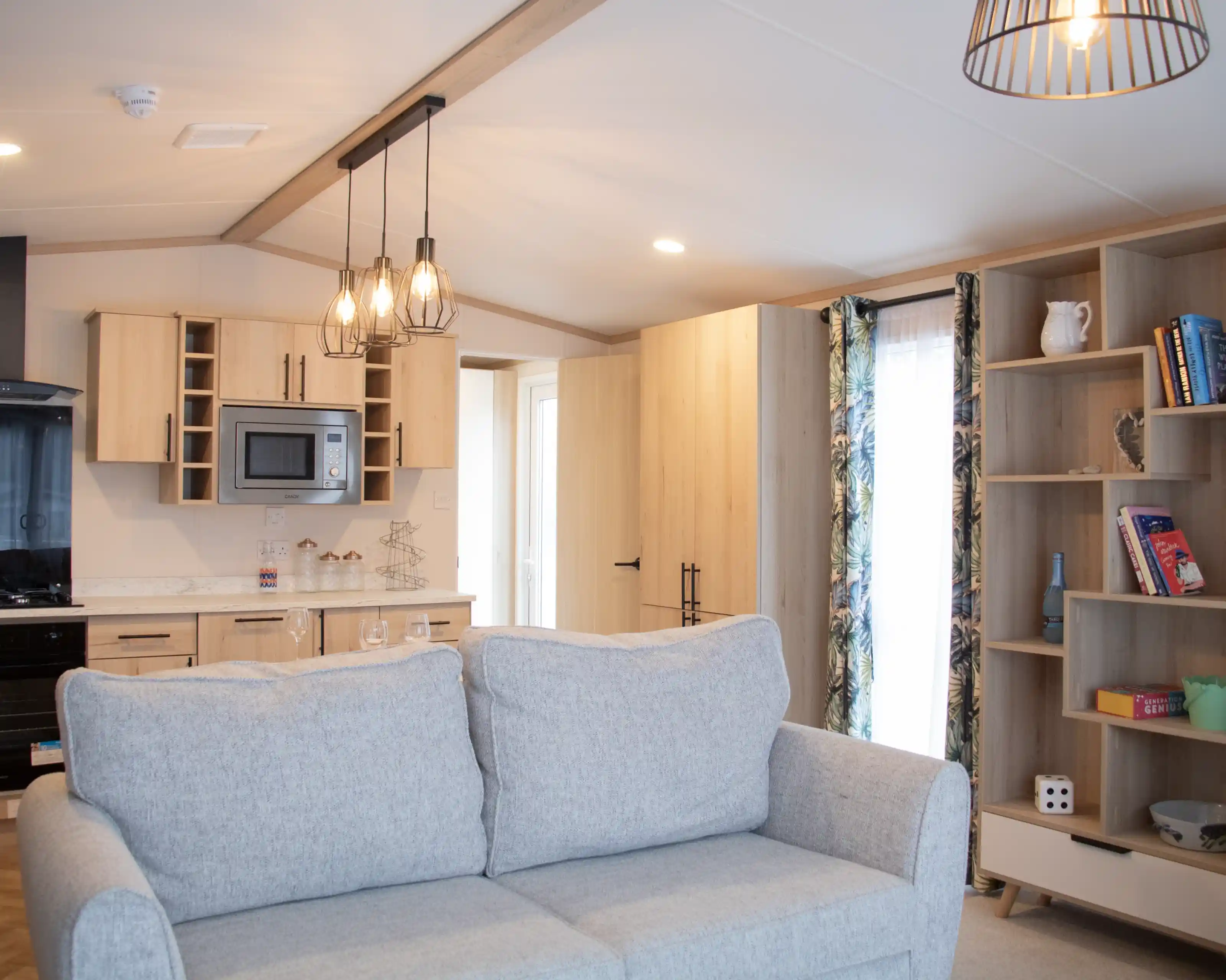 A cozy interior featuring a light gray couch in the foreground. In the background, a modern kitchen with wooden cabinetry, a microwave, and a dining area is visible. A shelf with books and decorative items is on the right, and a doorway leads to another room. The space is well-lit with a pendant light above.