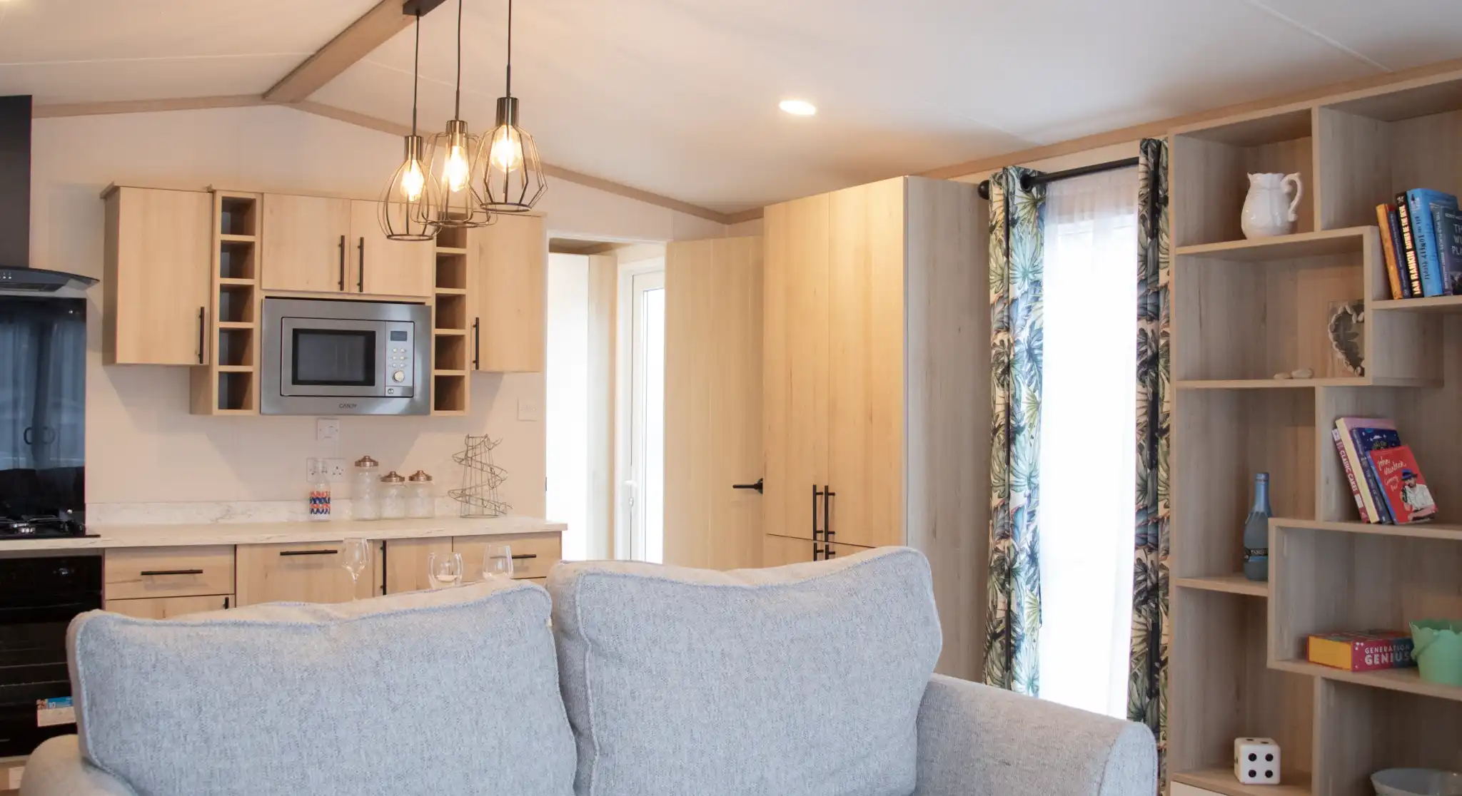 A cozy interior featuring a light gray couch in the foreground. In the background, a modern kitchen with wooden cabinetry, a microwave, and a dining area is visible. A shelf with books and decorative items is on the right, and a doorway leads to another room. The space is well-lit with a pendant light above.