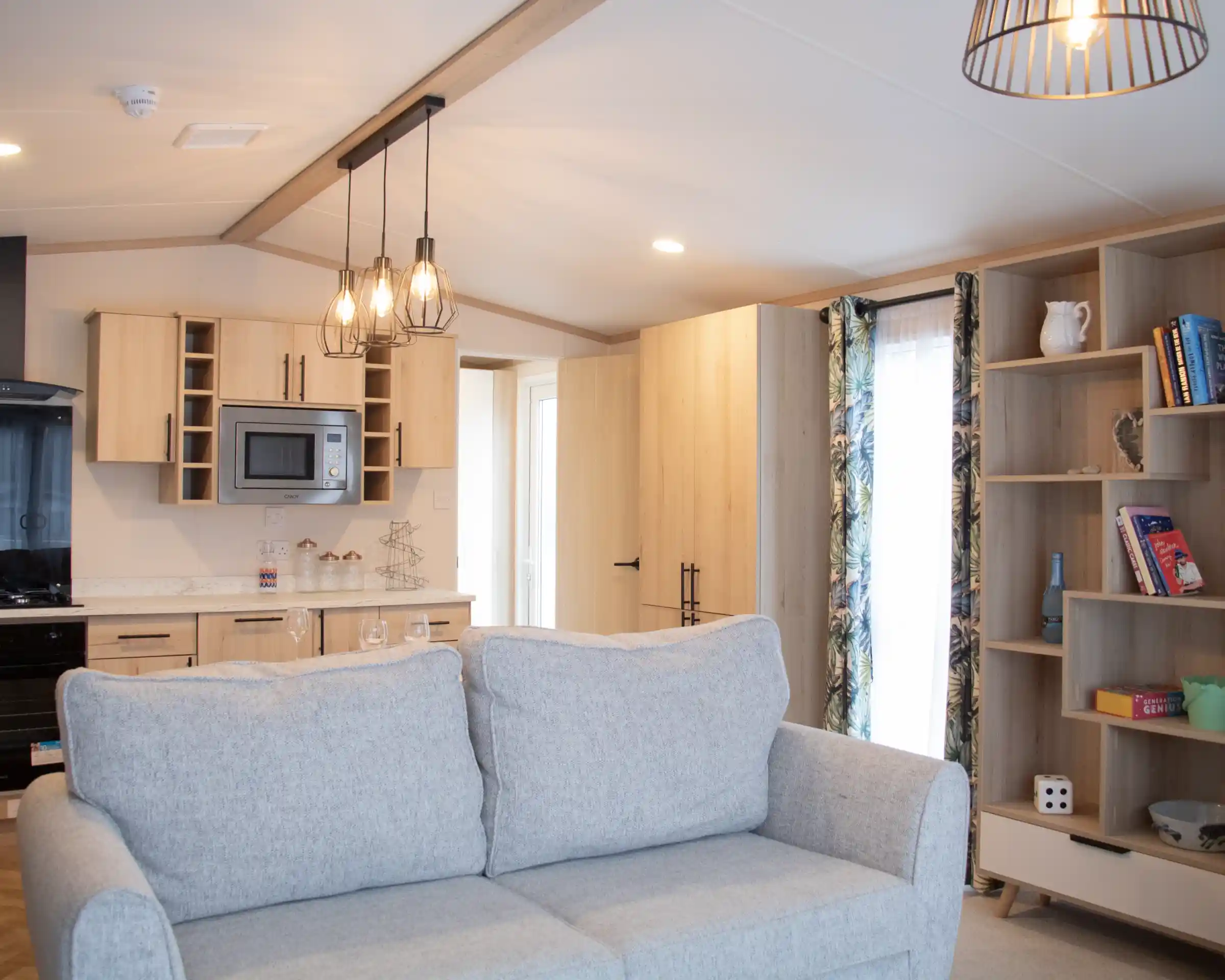 A cozy interior featuring a light gray couch in the foreground. In the background, a modern kitchen with wooden cabinetry, a microwave, and a dining area is visible. A shelf with books and decorative items is on the right, and a doorway leads to another room. The space is well-lit with a pendant light above.