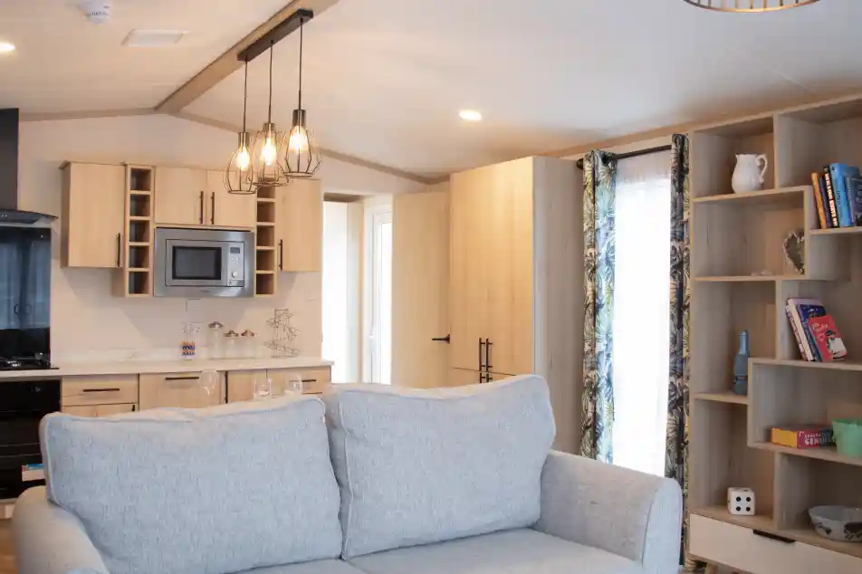 A cozy interior featuring a light gray couch in the foreground. In the background, a modern kitchen with wooden cabinetry, a microwave, and a dining area is visible. A shelf with books and decorative items is on the right, and a doorway leads to another room. The space is well-lit with a pendant light above.