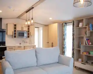 A cozy interior featuring a light gray couch in the foreground. In the background, a modern kitchen with wooden cabinetry, a microwave, and a dining area is visible. A shelf with books and decorative items is on the right, and a doorway leads to another room. The space is well-lit with a pendant light above.