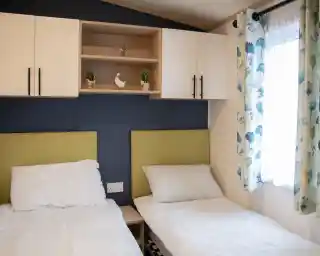 Two single beds with white bedding are positioned against a blue wall. Above the beds, a wooden shelf holds decorative plants and a small ornamental bird. A window with light curtains adorned with a floral pattern adds natural light to the room.