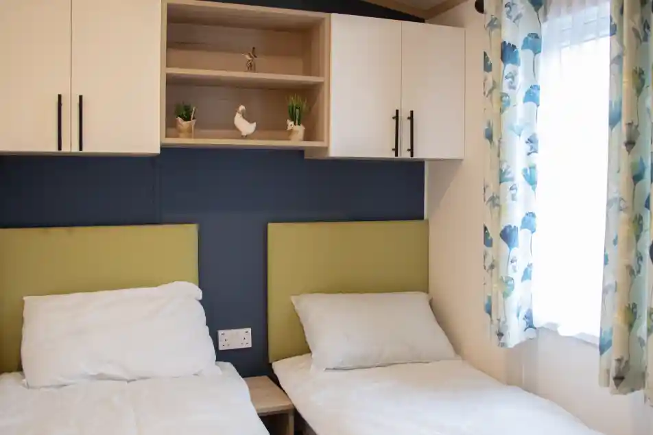 Two single beds with white bedding are positioned against a blue wall. Above the beds, a wooden shelf holds decorative plants and a small ornamental bird. A window with light curtains adorned with a floral pattern adds natural light to the room.