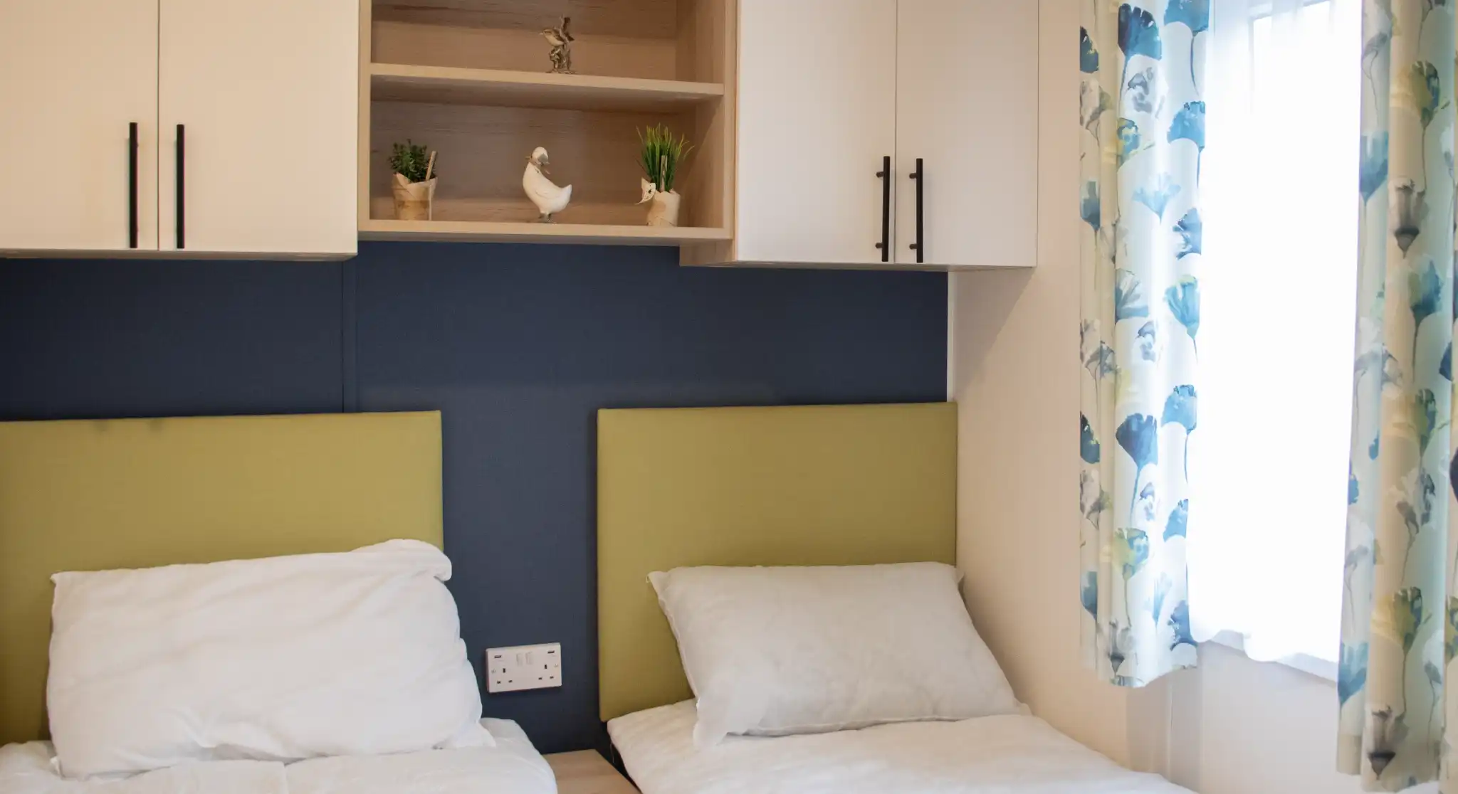Two single beds with white bedding are positioned against a blue wall. Above the beds, a wooden shelf holds decorative plants and a small ornamental bird. A window with light curtains adorned with a floral pattern adds natural light to the room.