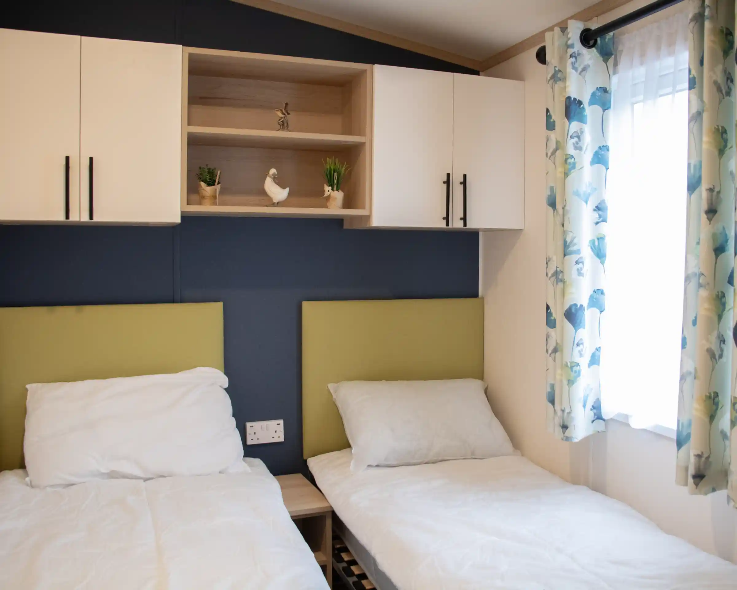 Two single beds with white bedding are positioned against a blue wall. Above the beds, a wooden shelf holds decorative plants and a small ornamental bird. A window with light curtains adorned with a floral pattern adds natural light to the room.